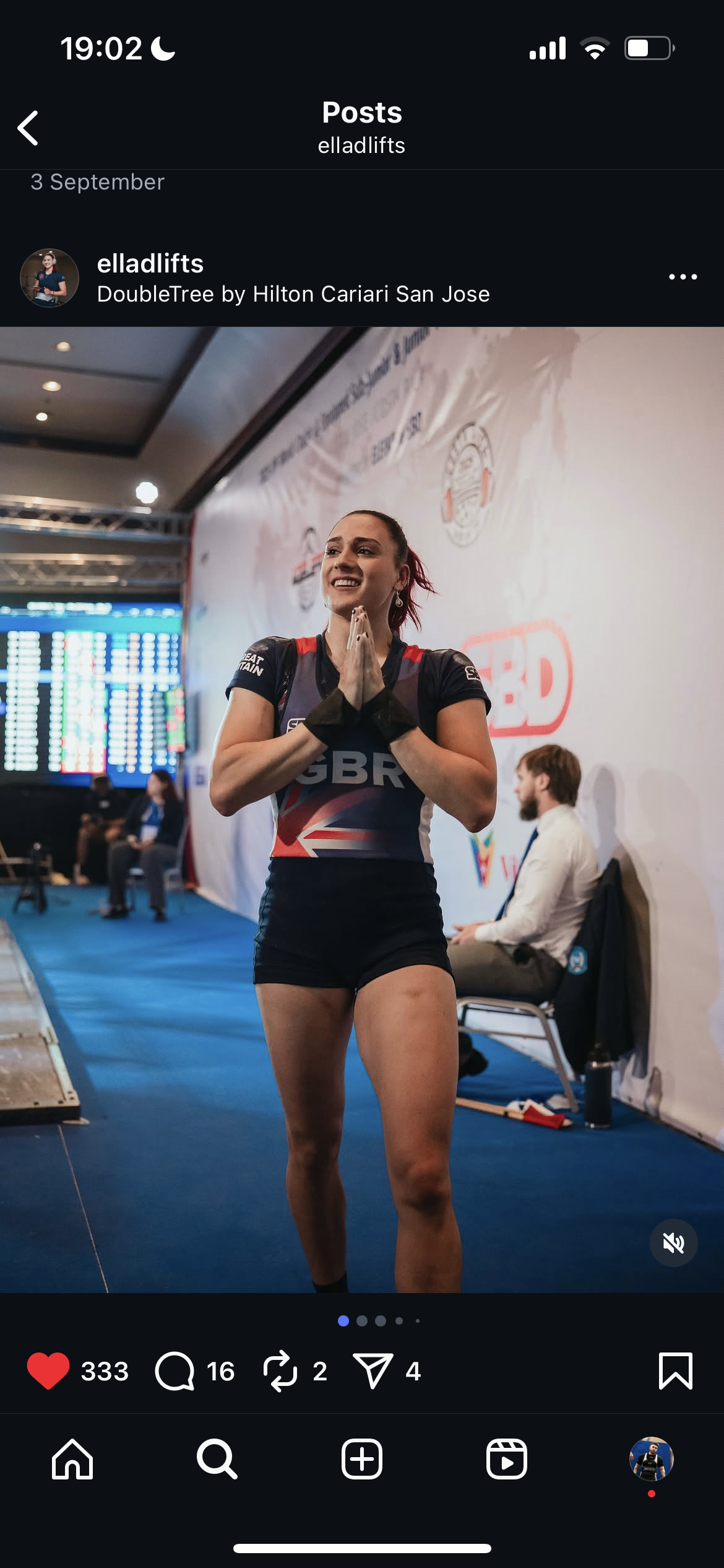 A female athlete, wearing a black athletic uniform with 'GBR' and a Union Jack flag, smiling with hands clasped together, standing in an indoor sports arena.