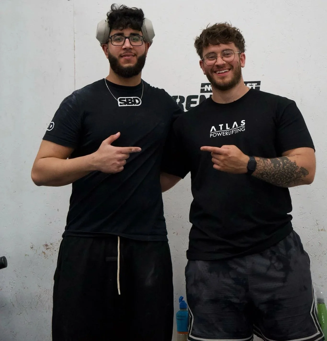 Two men smiling at the camera, pointing at each other, standing against a plain white wall. The man on the left wears headphones around his head, glasses, a black T-shirt, and black shorts. The man on the right wears glasses, a black T-shirt with 'ATLAS POWERLIFTING' printed on it, and camouflage shorts.