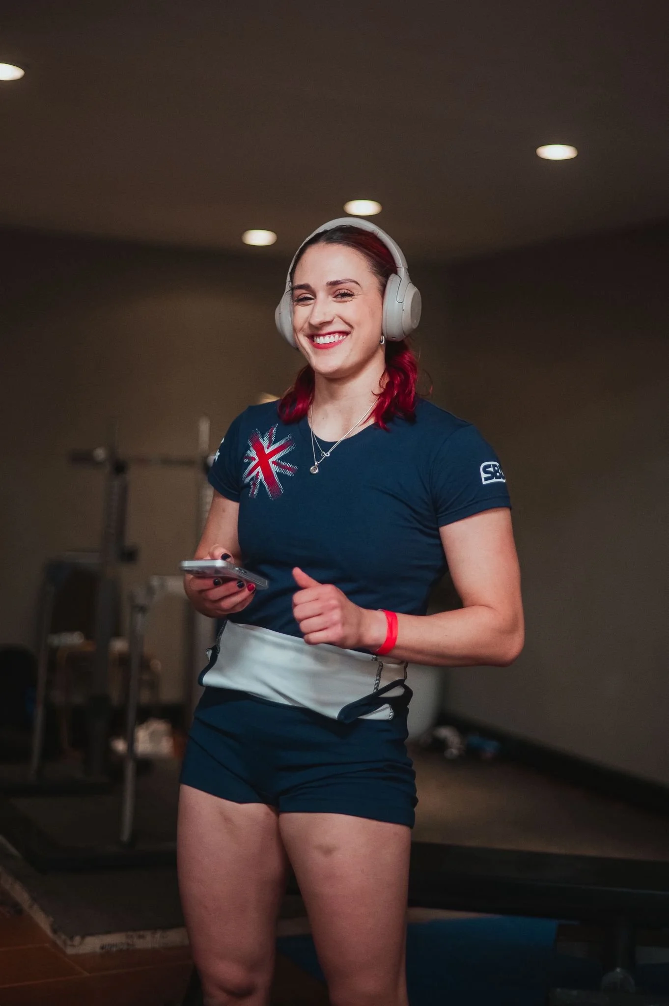 A young woman with red hair smiling, wearing headphones, a navy T-shirt with a British flag, and athletic shorts, standing in a gym.