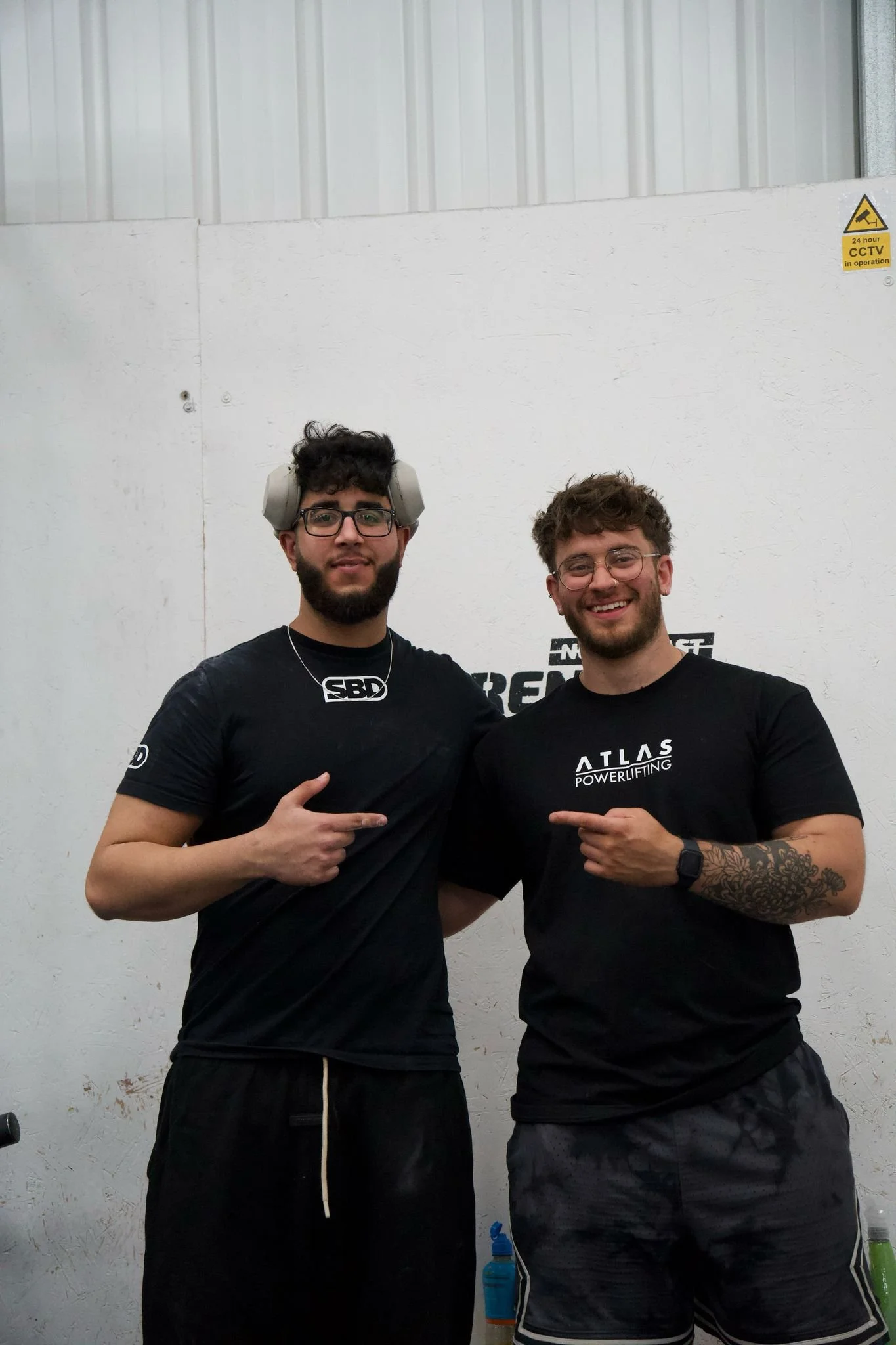 Two young men standing together in a gym, smiling and pointing at each other. One wears headphones and glasses, the other has tattoos on his arm and is wearing a smartwatch. They are dressed in black athletic clothing.