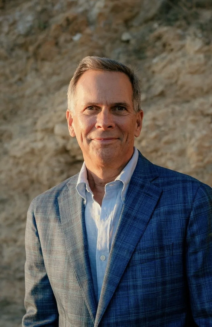 A middle-aged man in a blue plaid blazer and white shirt posing outdoors with a rocky background.