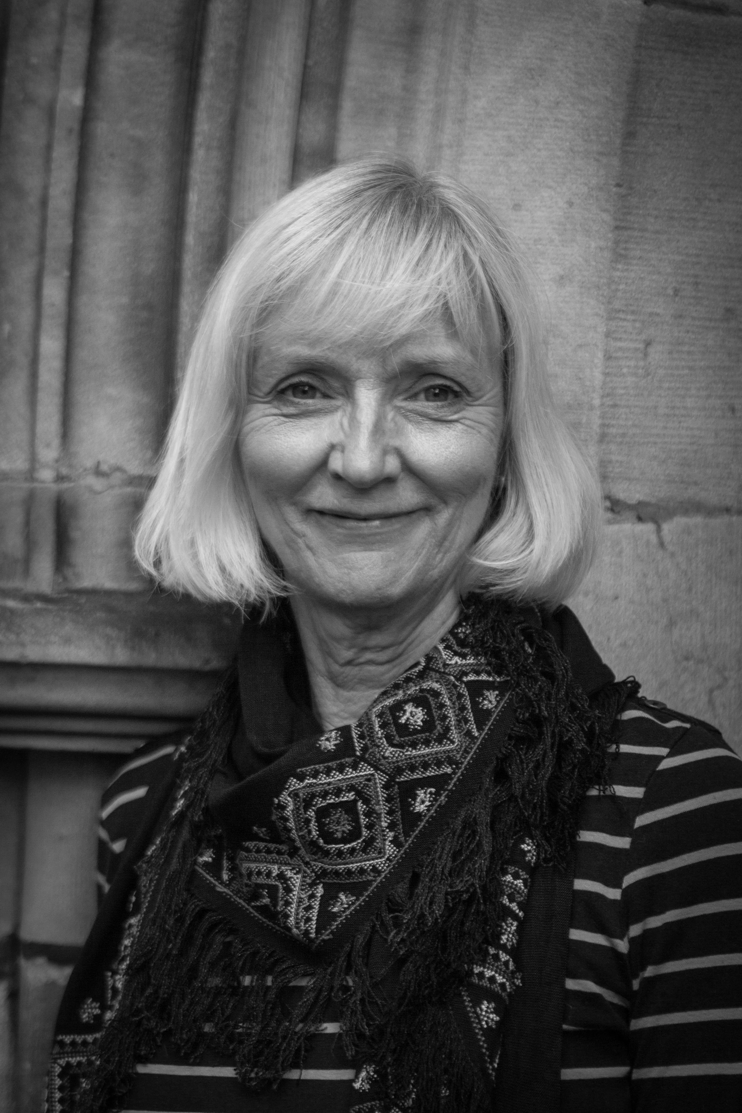 A black and white photo of a smiling middle-aged woman with shoulder-length blonde hair, wearing a striped shirt and an embroidered scarf, standing in front of a stone wall with curtains.