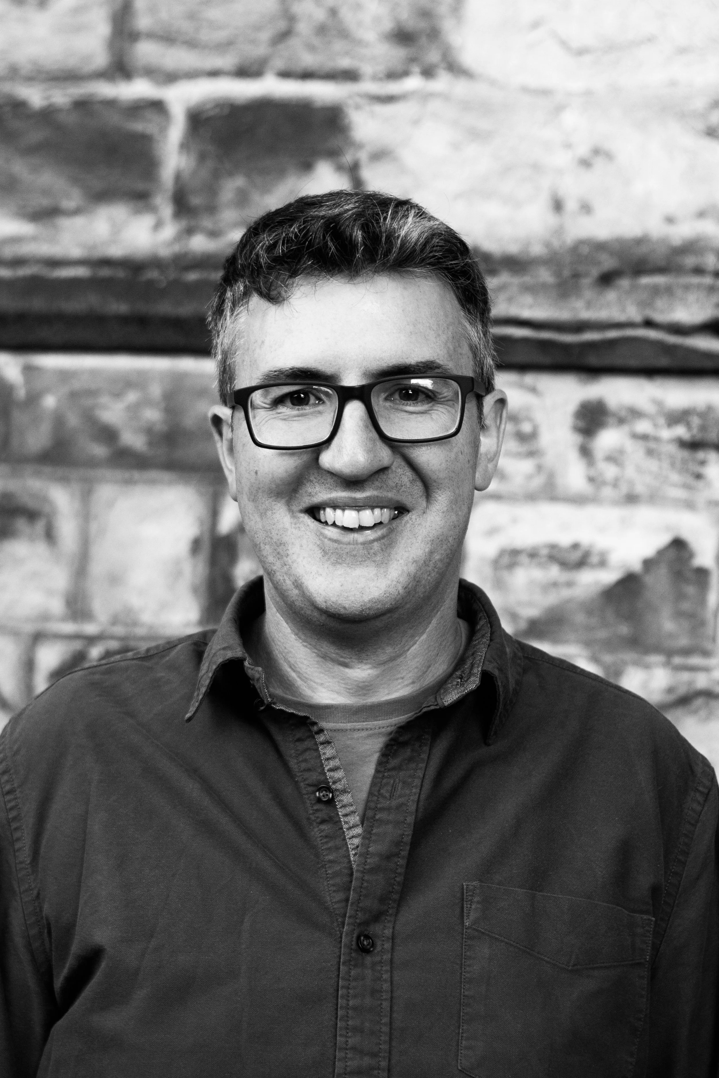 Black and white portrait of a smiling man wearing glasses and a button-up shirt in front of a stone wall.