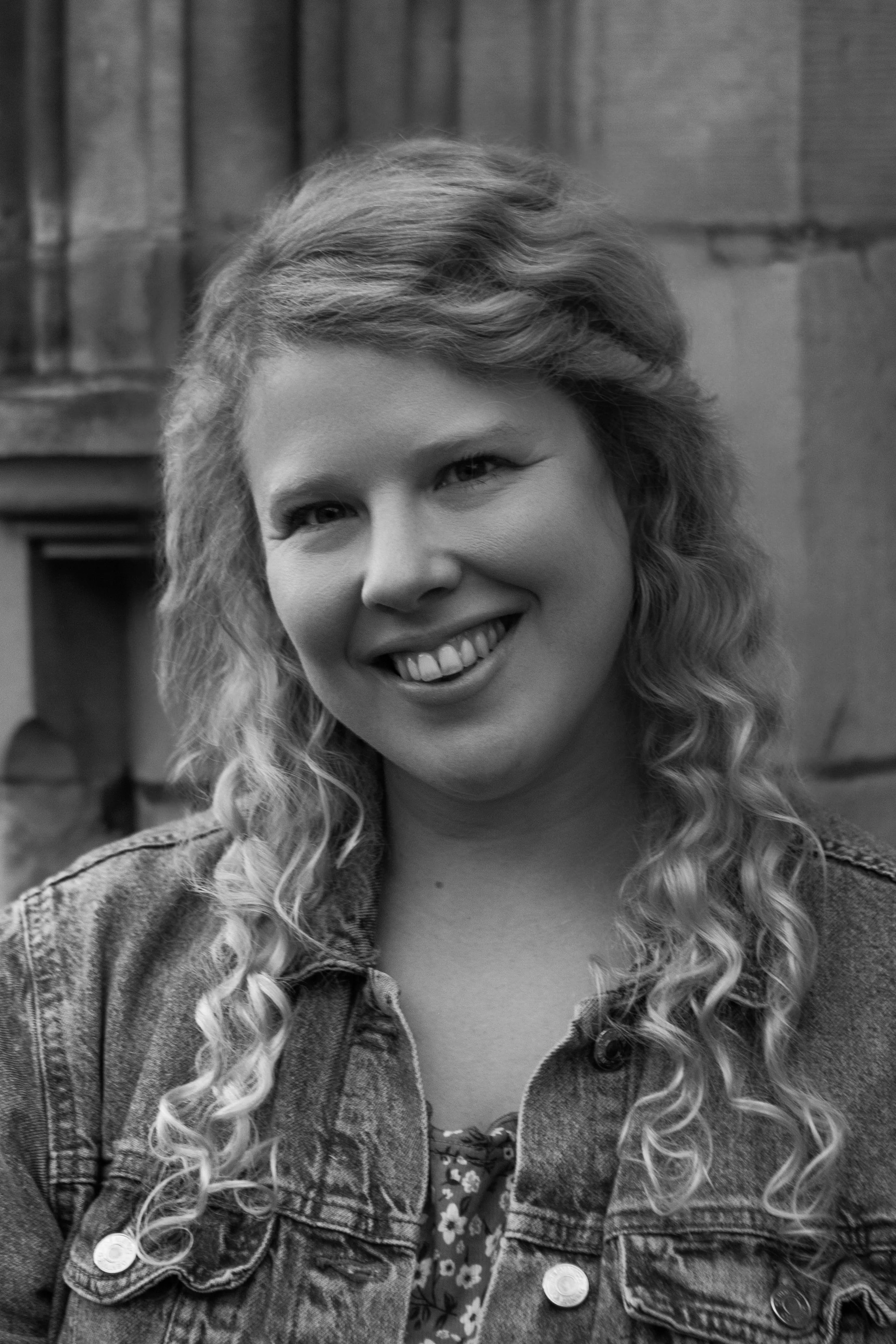 Black and white portrait of a smiling woman with long, curly hair wearing a textured coat and a buttoned shirt, with a stone or brick wall in the background.