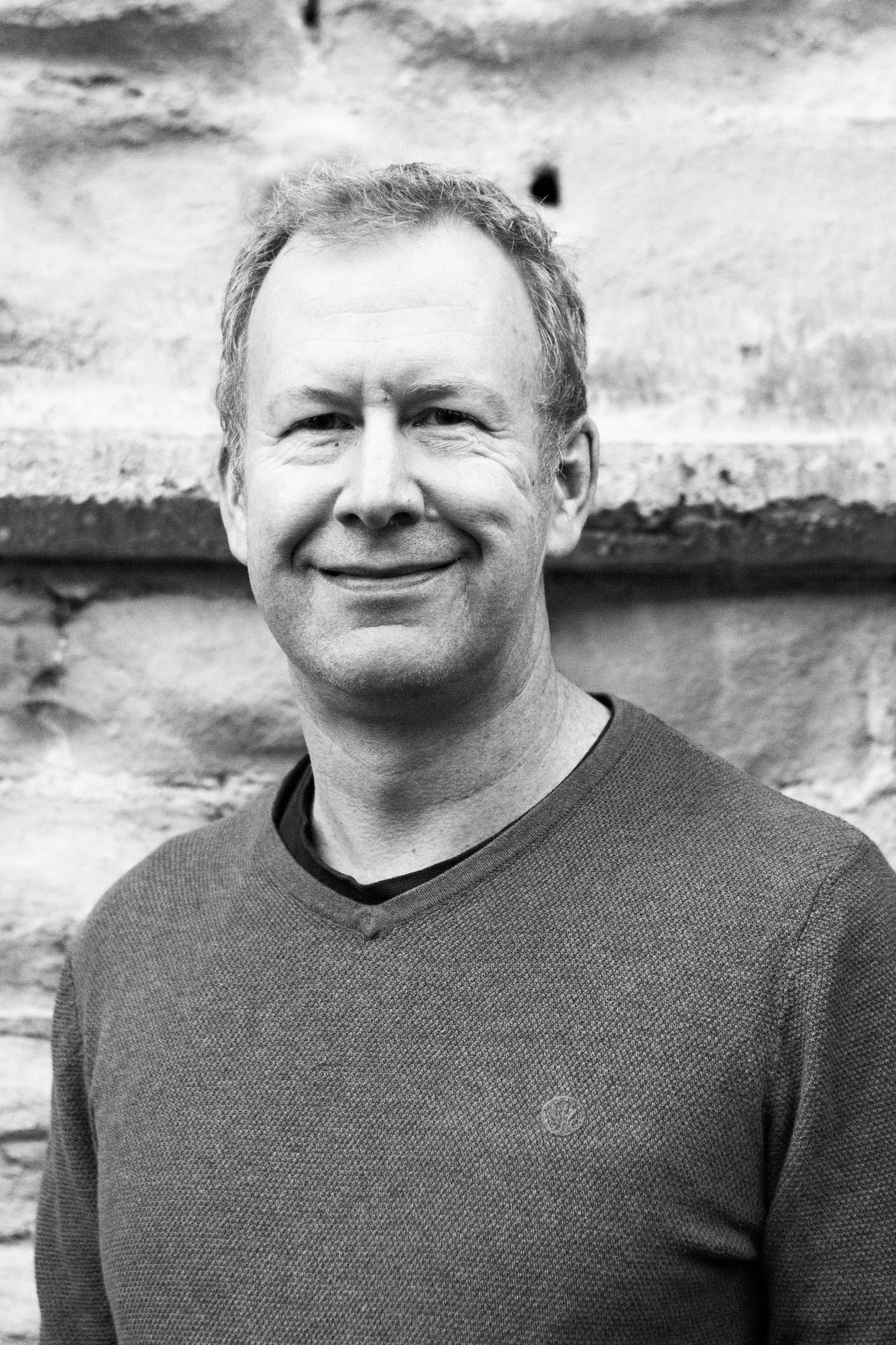 Black and white portrait of a smiling man standing in front of a brick wall.