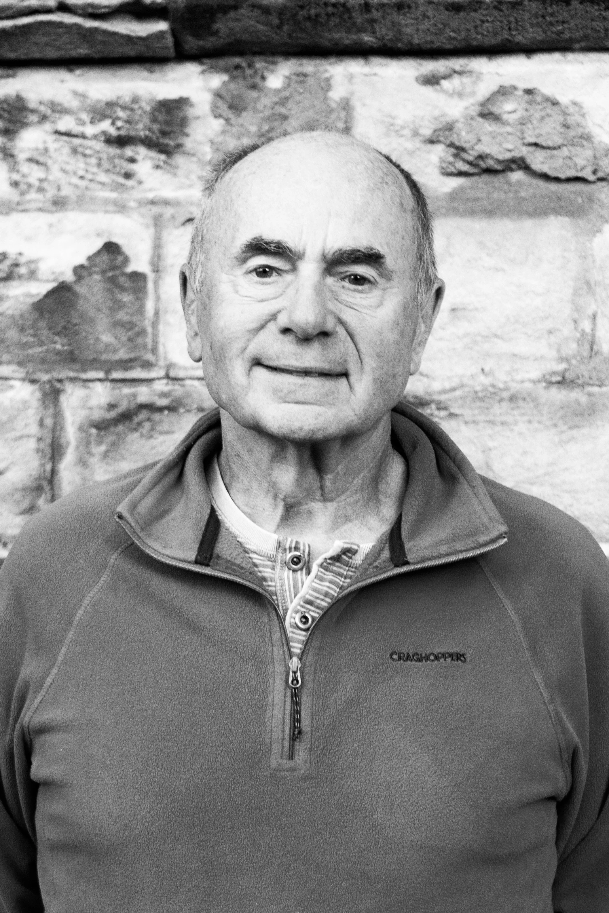 A black and white photo of an elderly man with a bald head and prominent eyebrows, wearing a zip-up jacket over a striped shirt, standing in front of a stone wall.