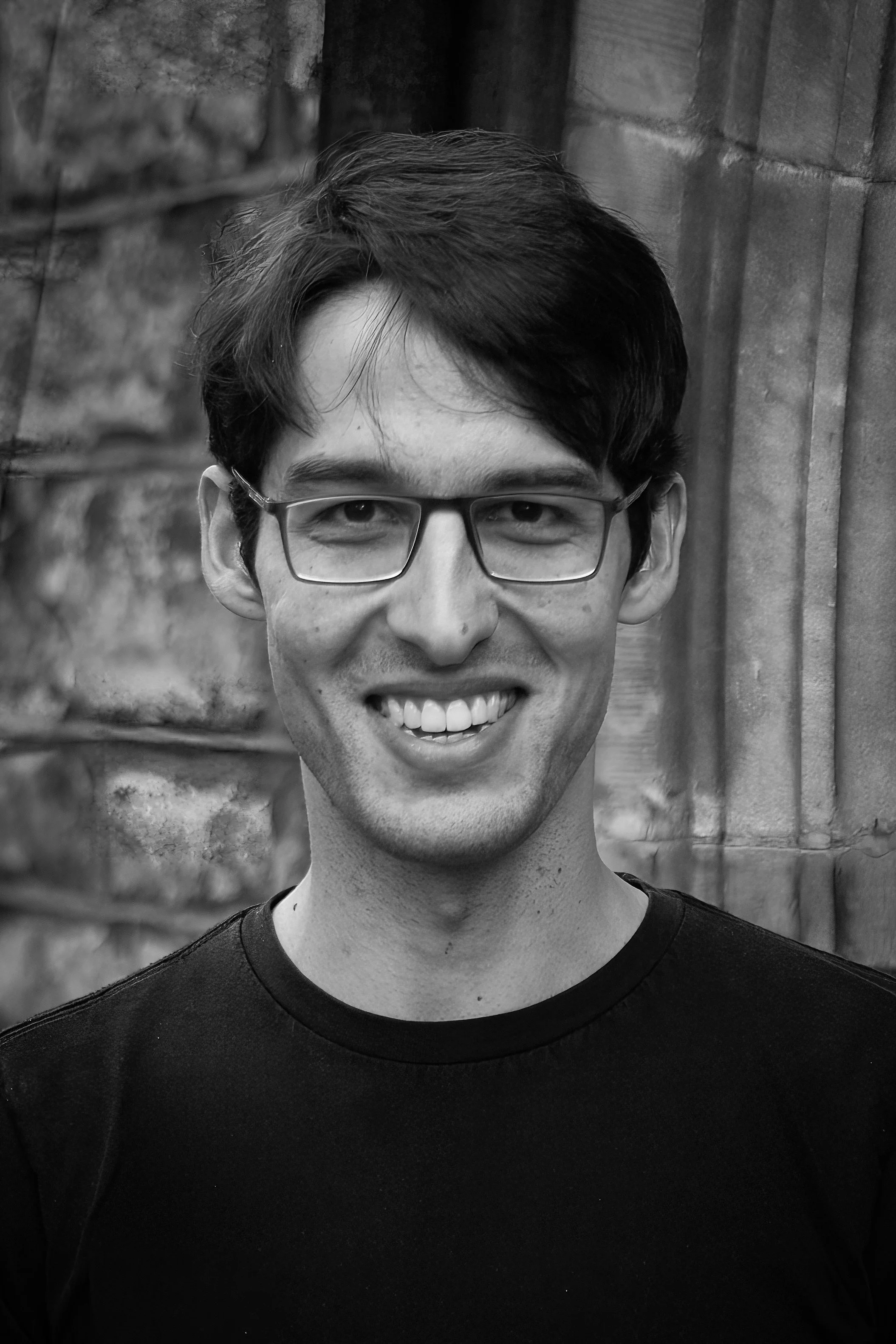Black and white portrait of a smiling man with glasses, wearing a black shirt, standing outdoors in front of a stone wall.