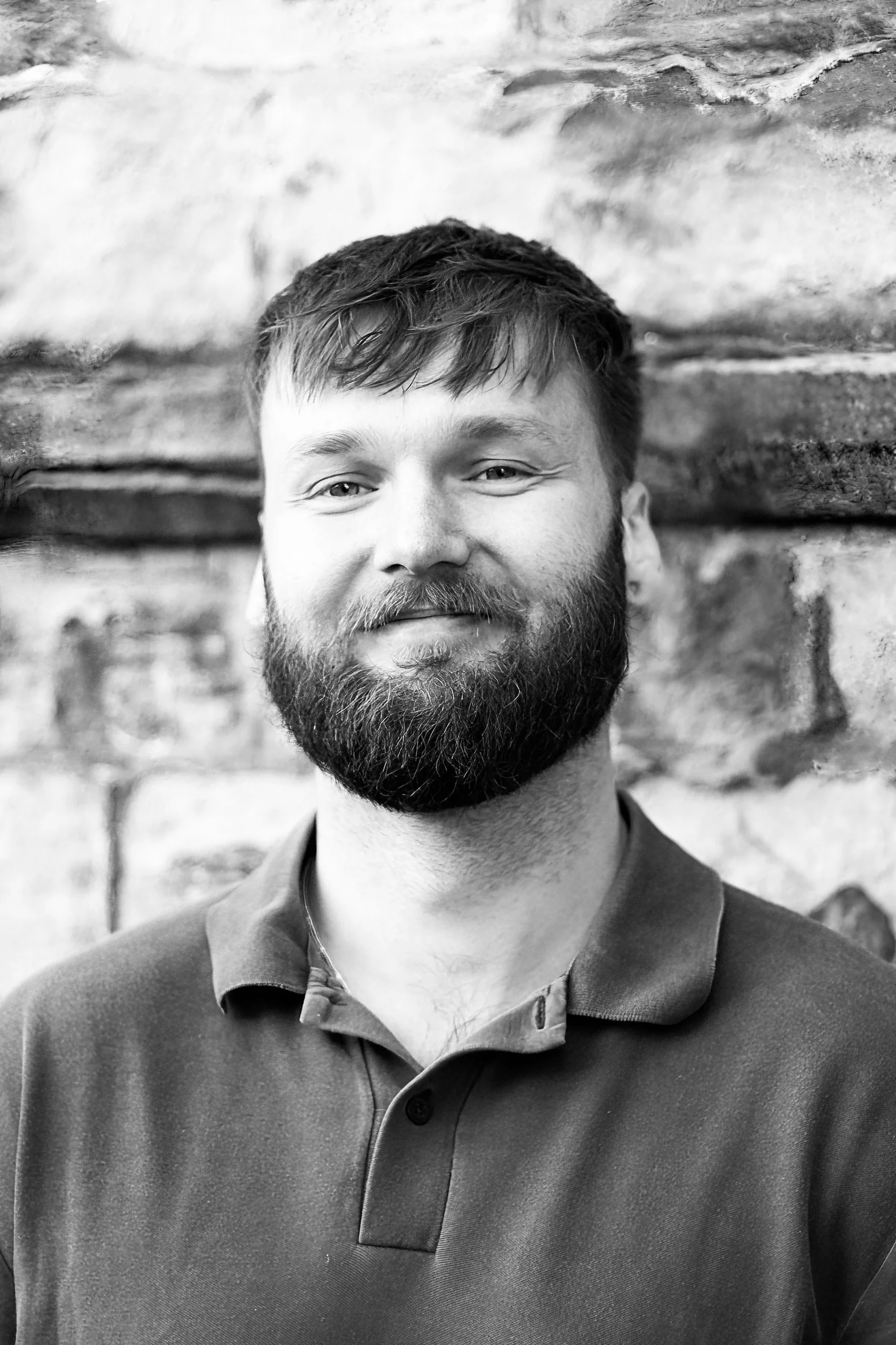 A black and white portrait of a smiling man with a beard and short hair, wearing a polo shirt, standing in front of a brick wall.