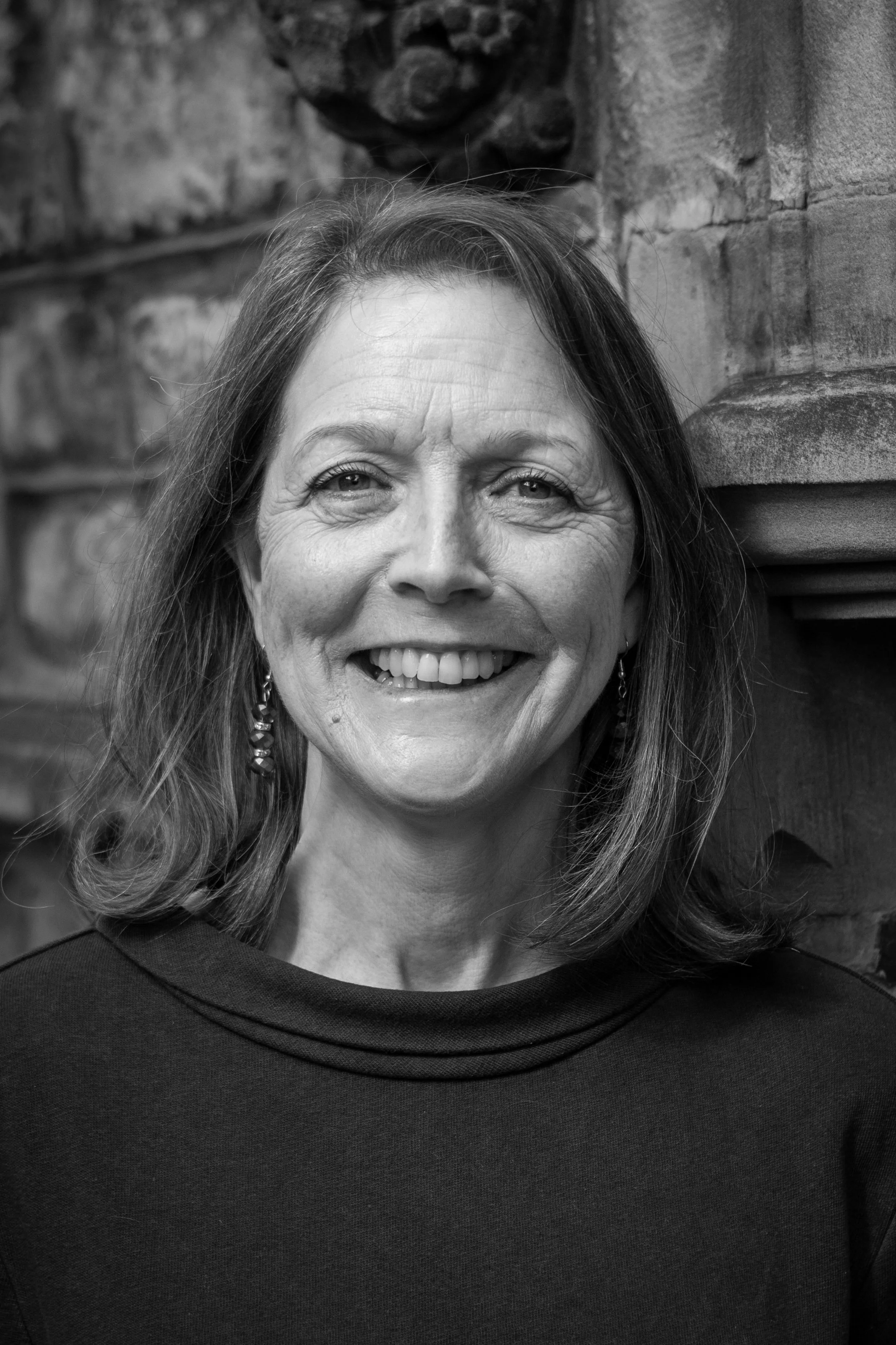 A black and white portrait of a smiling middle-aged woman with shoulder-length hair, wearing earrings and a dark top, standing in front of a brick and stone wall.