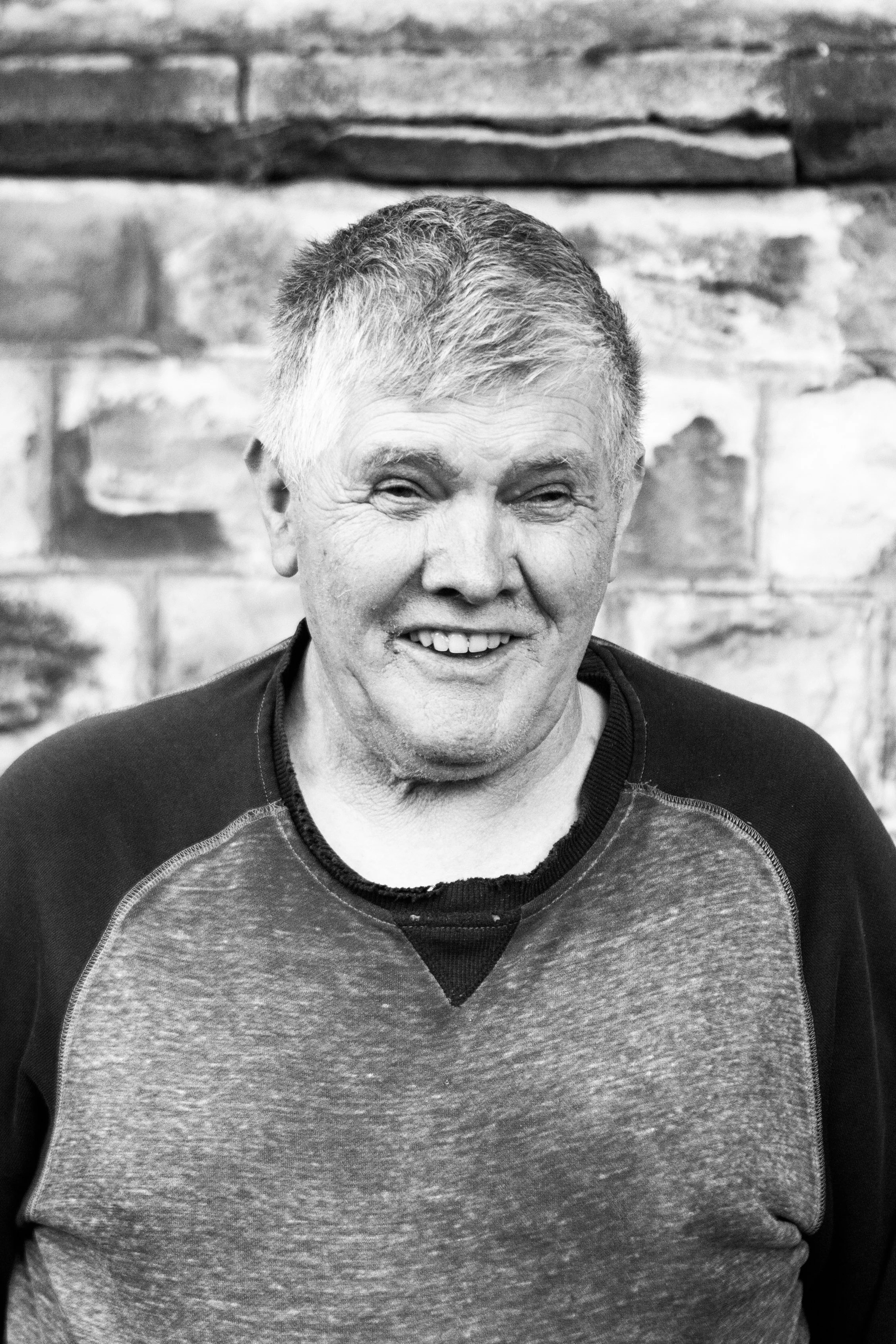 A smiling middle-aged man with short hair, wearing a casual long-sleeve shirt, standing against a brick wall.