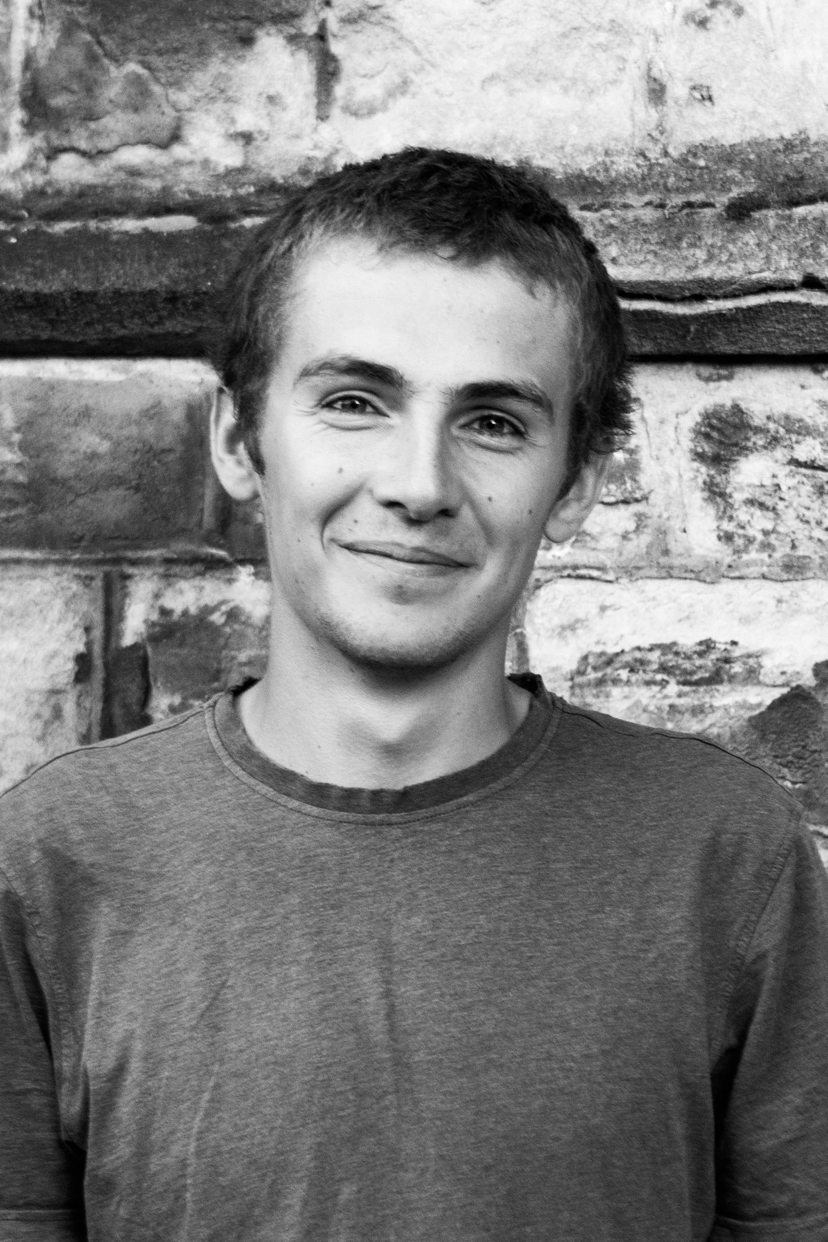 Black and white portrait of a young man with short hair smiling, standing in front of a brick wall.