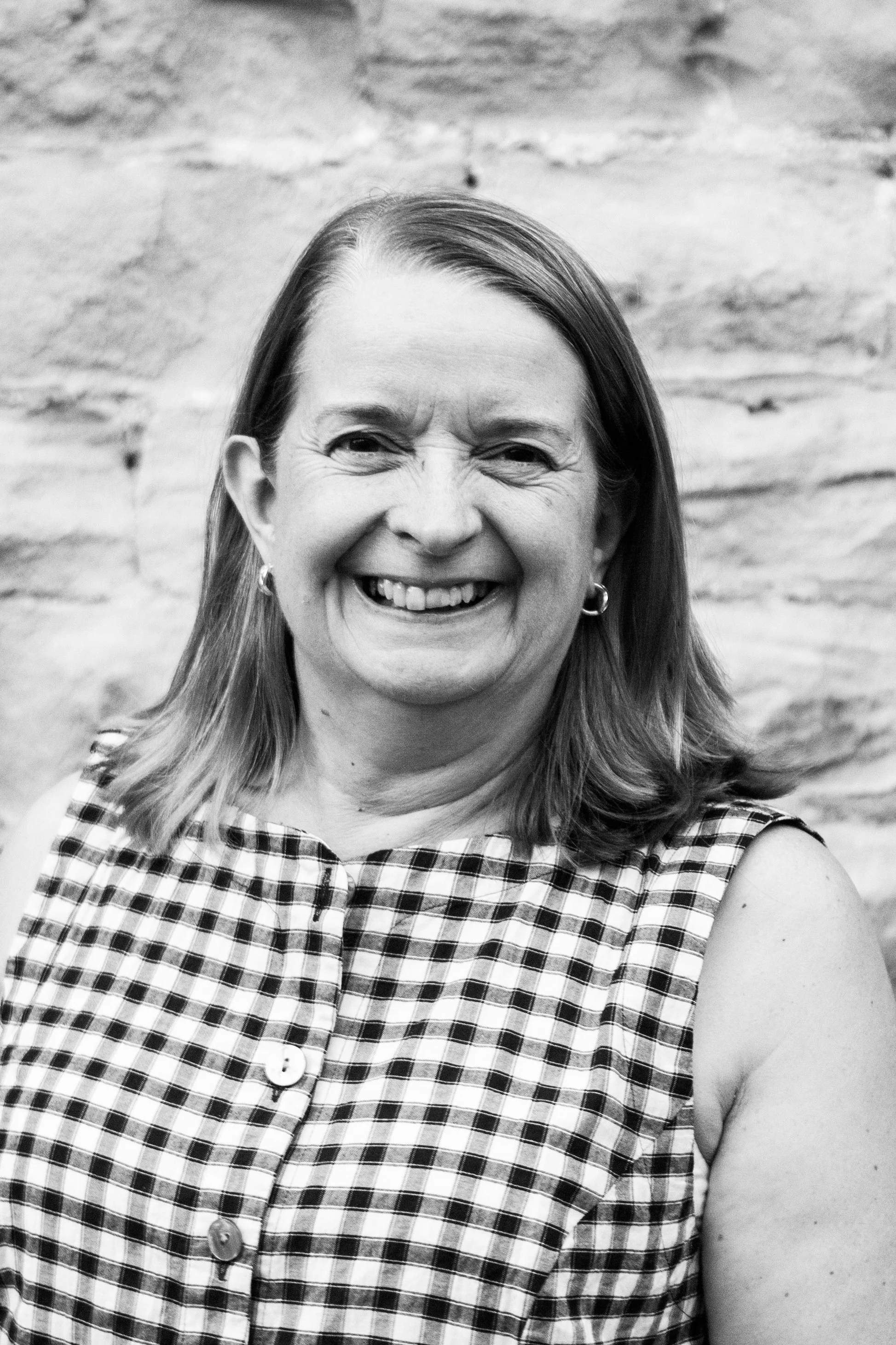 A smiling woman with shoulder-length hair, wearing a sleeveless checkered top, standing in front of a brick wall.