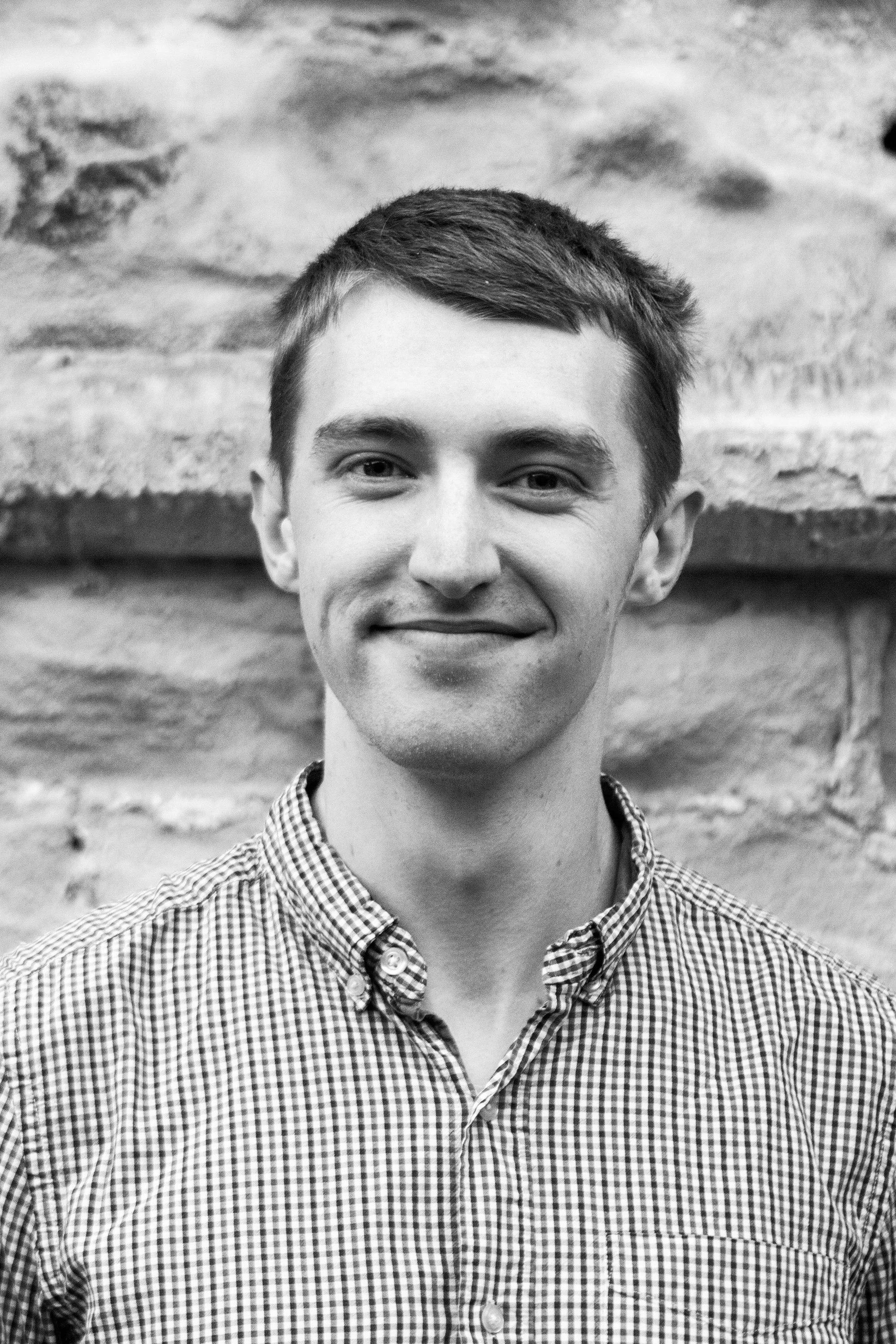 Black and white photo of a young man with short dark hair, smiling, wearing a checkered button-up shirt, standing in front of a textured brick wall.