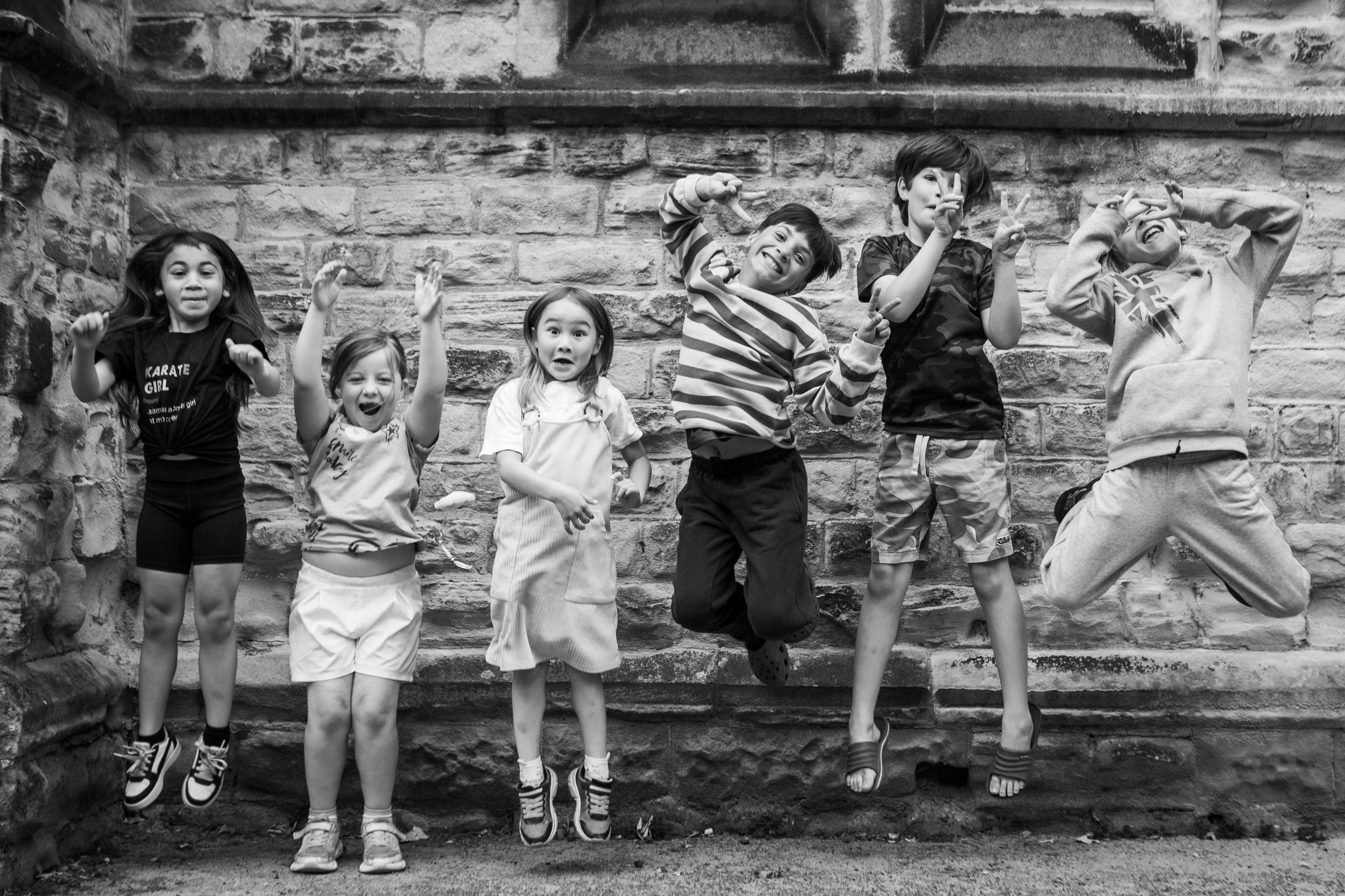Six children jumping and smiling in front of a brick wall, captured in black and white.