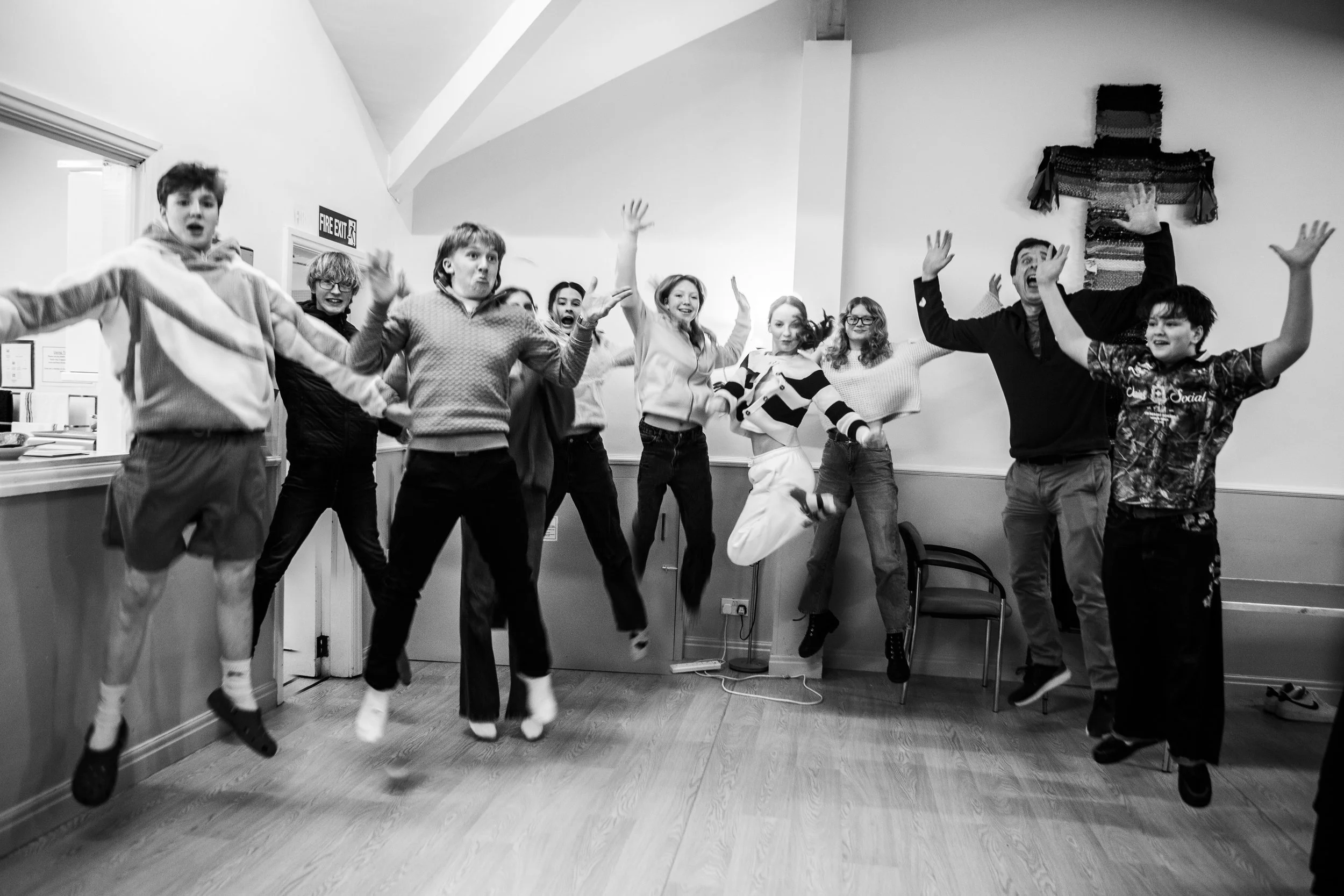Group of people in mid-air jump, smiling, in a room with hardwood floor and plain walls, celebrating or having fun.