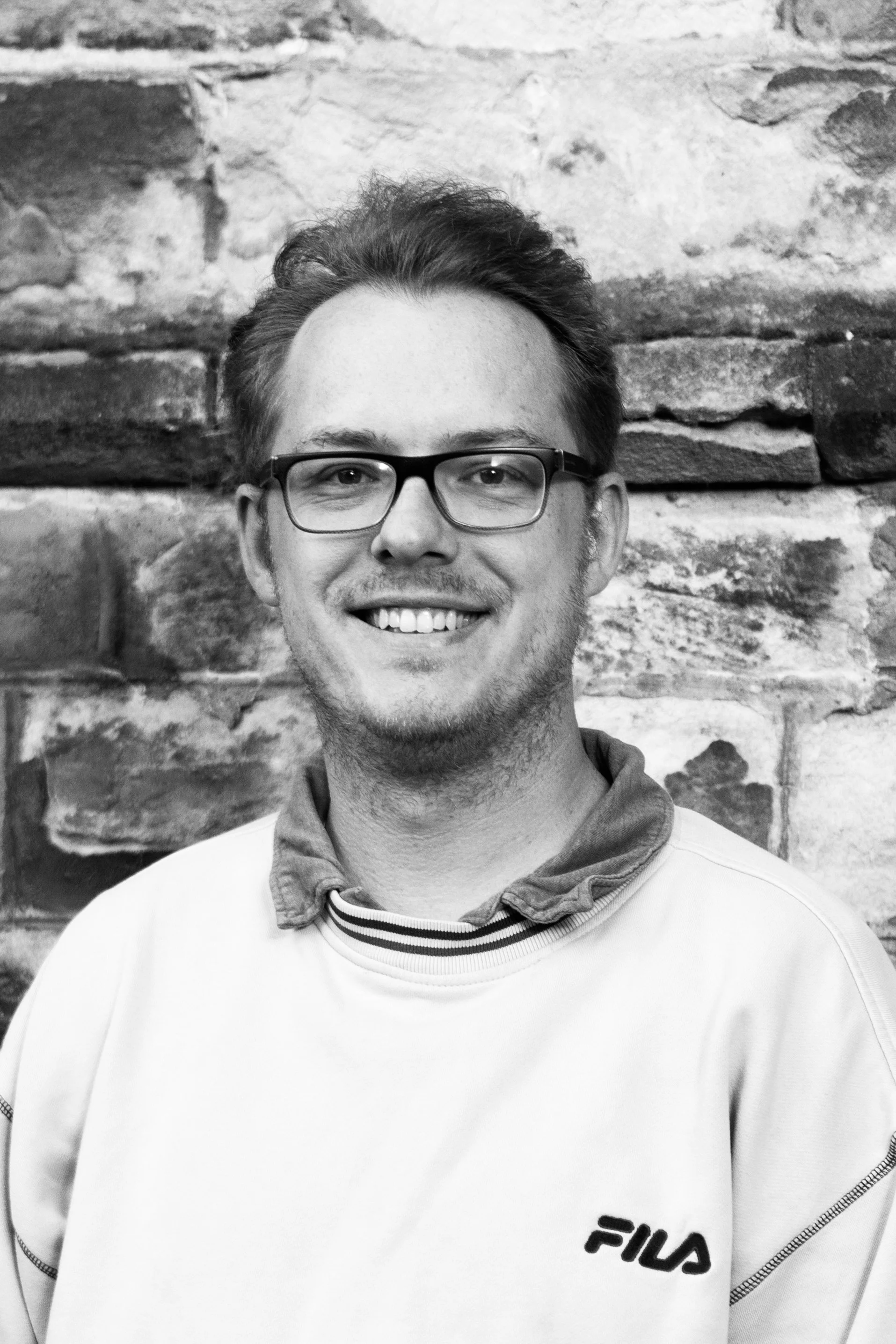 A young man with glasses smiling, wearing a FILA sweatshirt, standing in front of a brick wall.