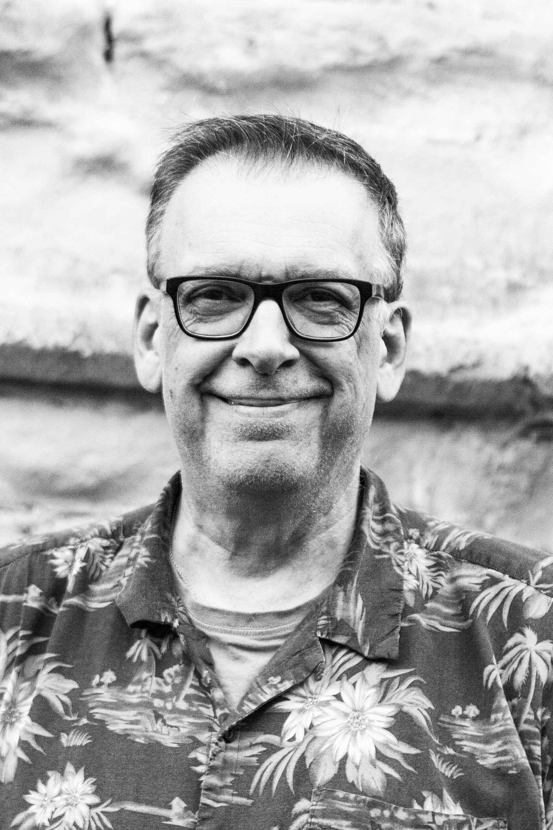 Black and white photo of a smiling man wearing glasses and a tropical patterned shirt, standing outdoors with a rocky background.