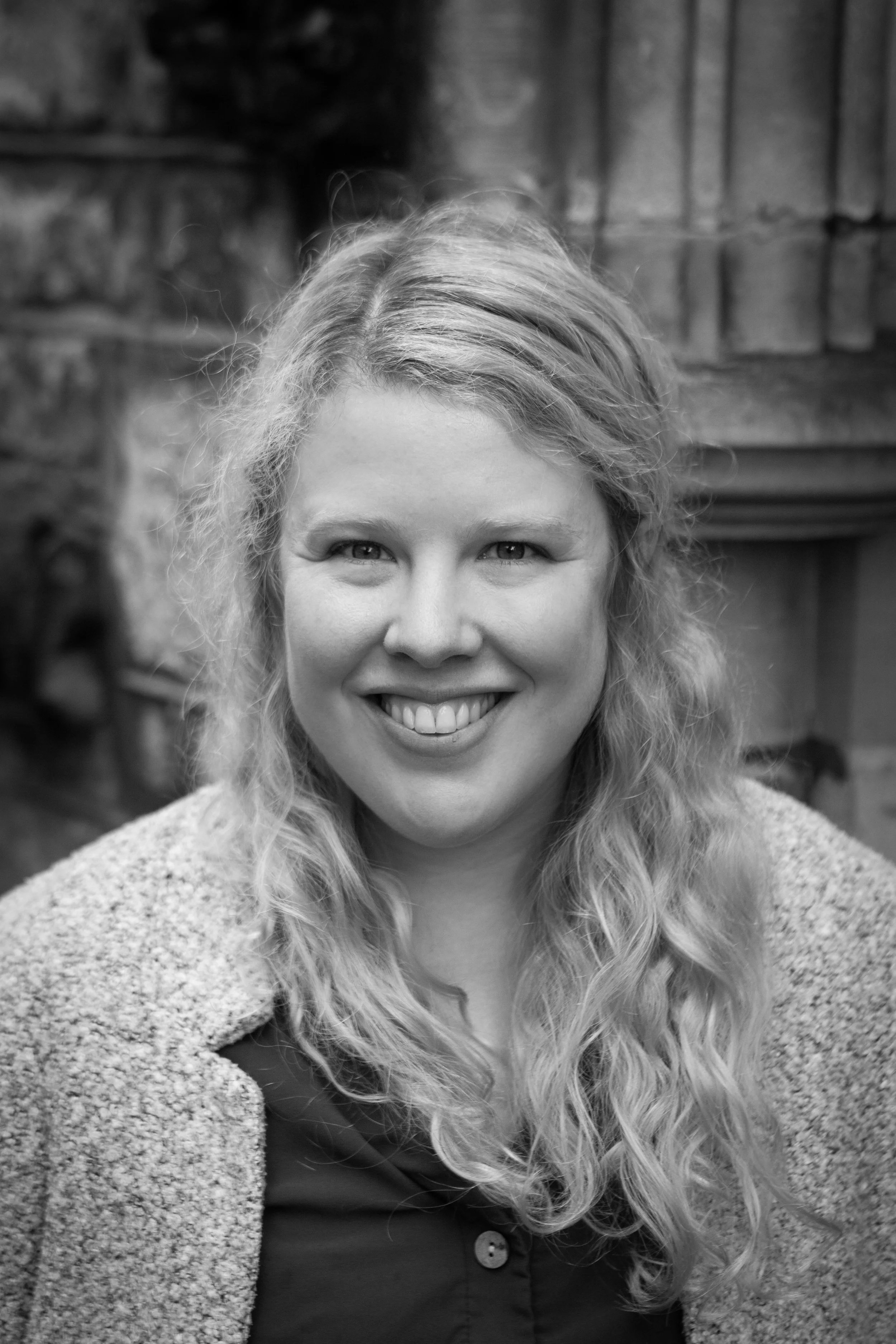 Black and white portrait of a smiling woman with long, curly hair wearing a textured coat and a buttoned shirt, with a stone or brick wall in the background.