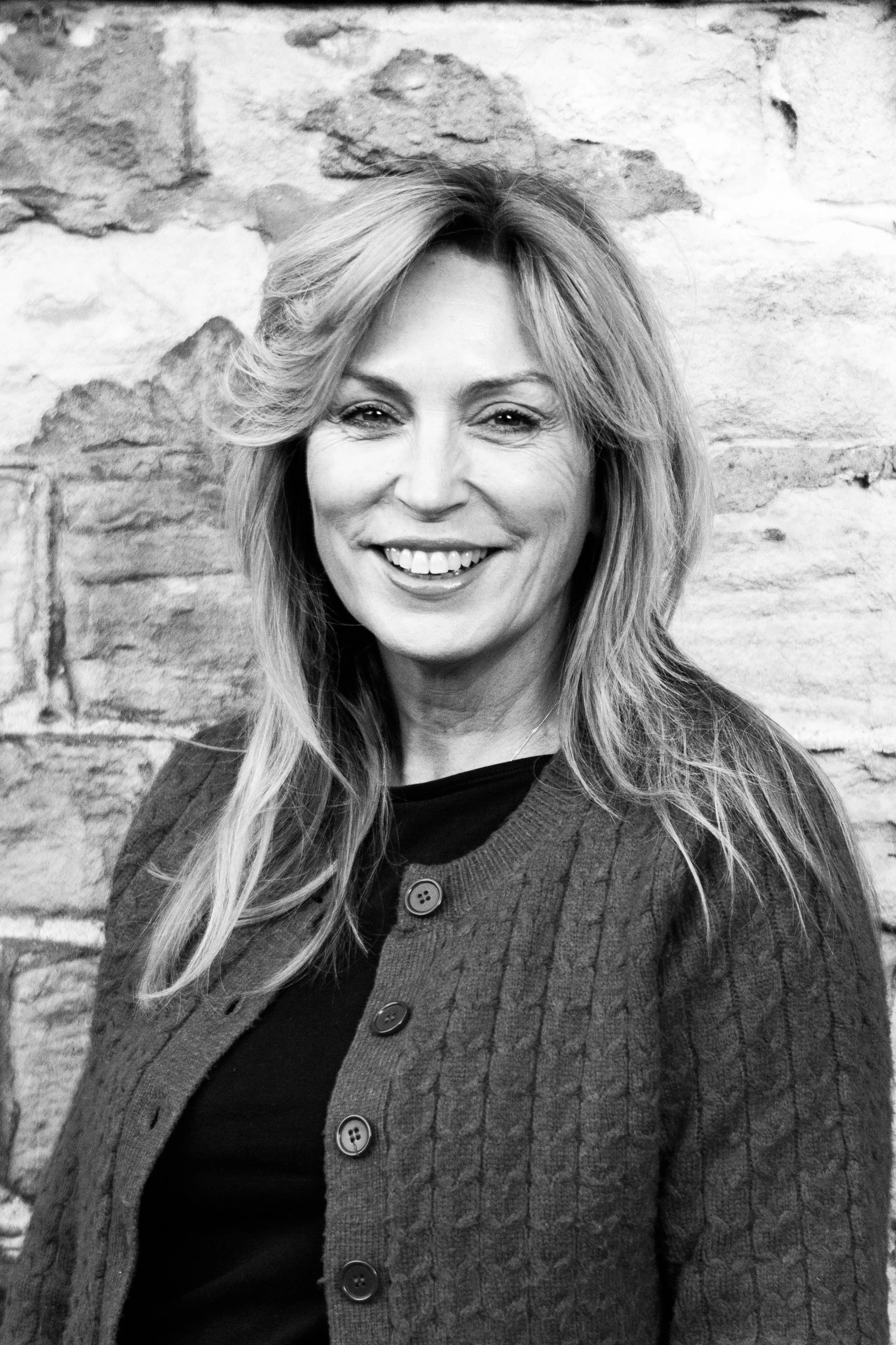 Black and white portrait of a smiling woman with shoulder-length hair, wearing a textured cardigan over a dark top, standing against a brick wall.