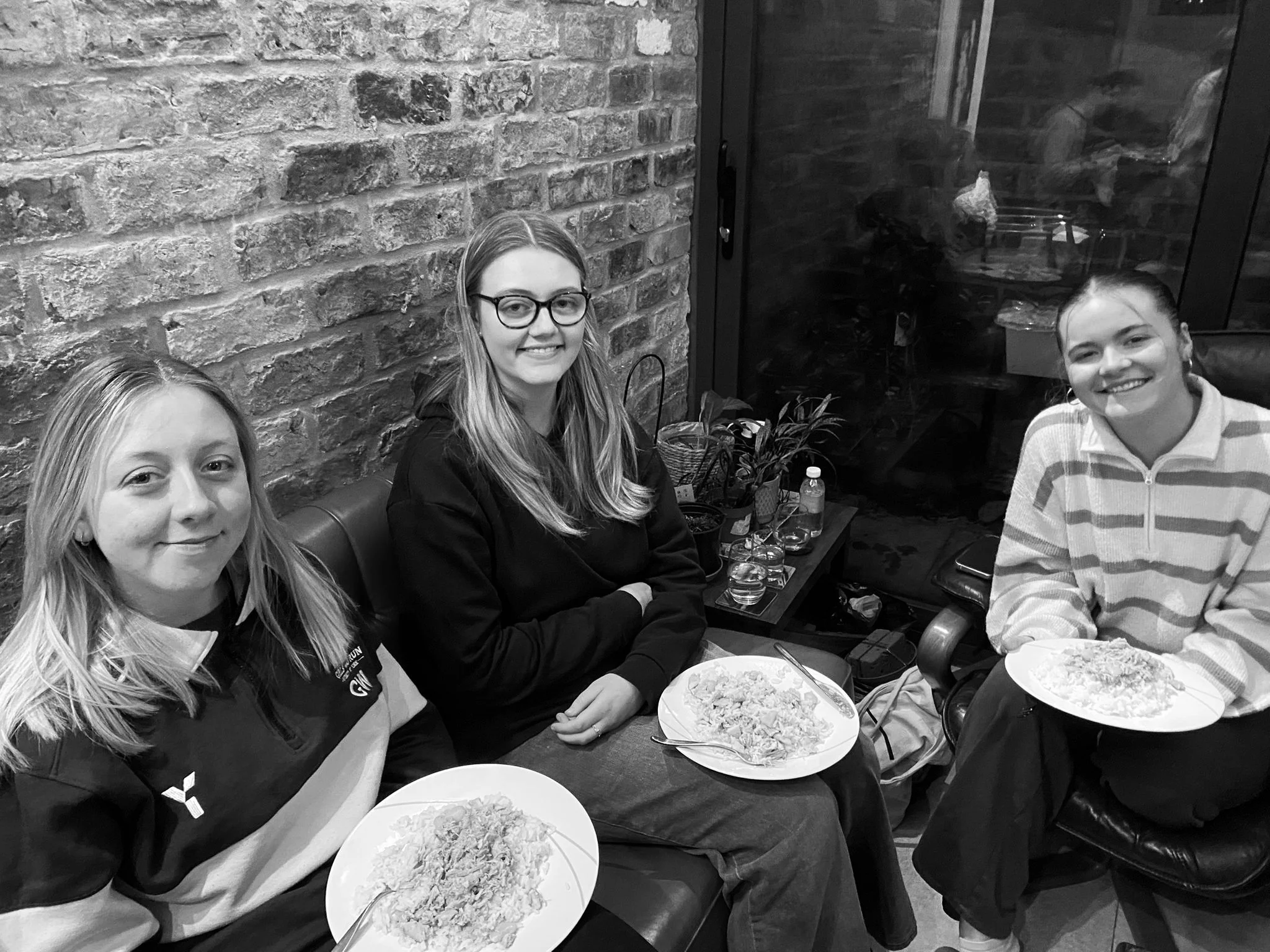 Three women sitting on a couch at a table with plates of food, smiling at the camera in a cozy indoor setting with a brick wall and large window behind them.
