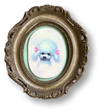 A small dog with pink blush marks and a pink bow on its head inside a decorative oval mirror frame.