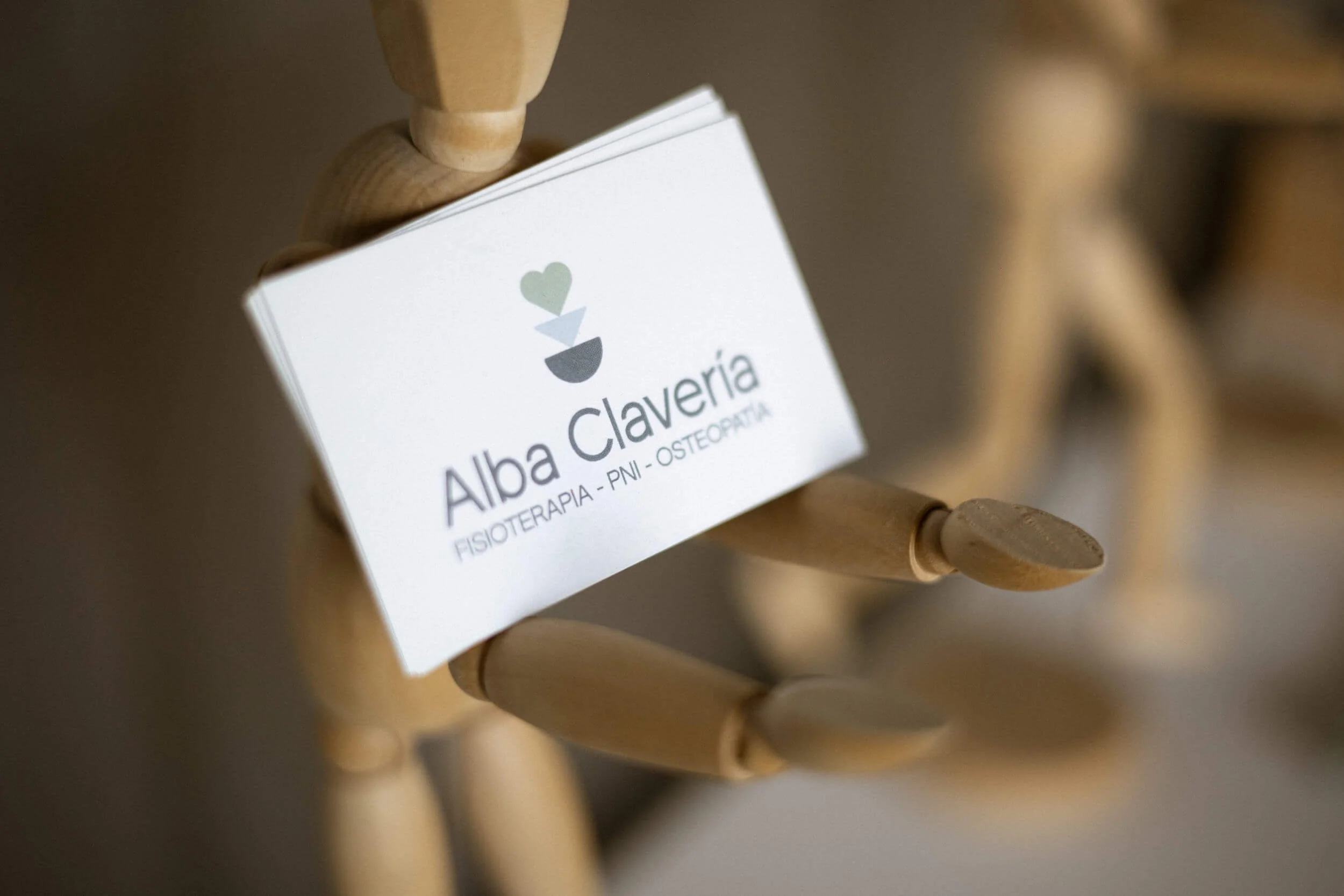 Tarjeta de presentación en una figura de madera que dice 'Alba Clavería, Fisioterapia, PNI, Osteopatía' con un ícono de un corazón.