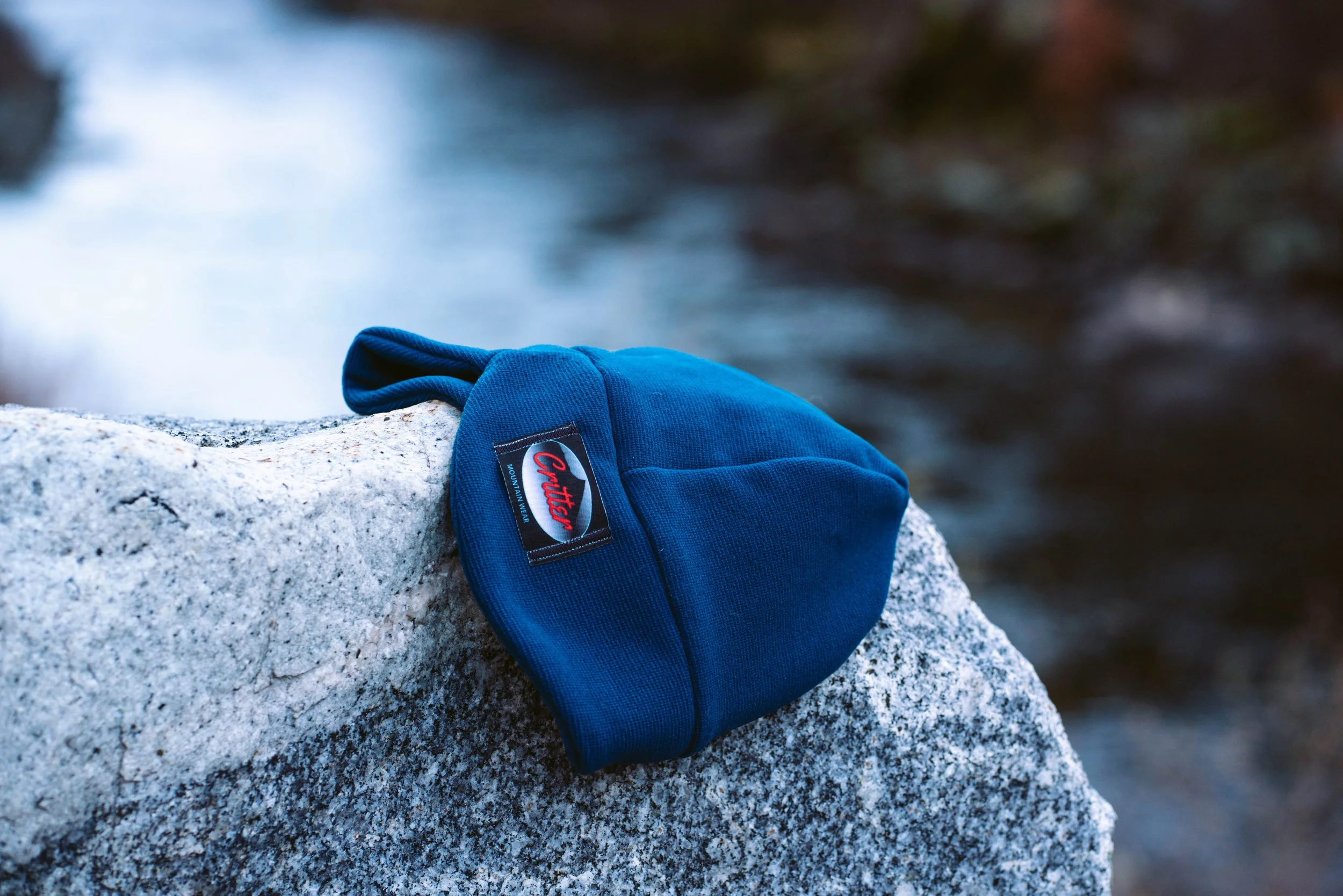 A blue Critter Mountain Wear hat resting on a large gray rock beside a flowing river.