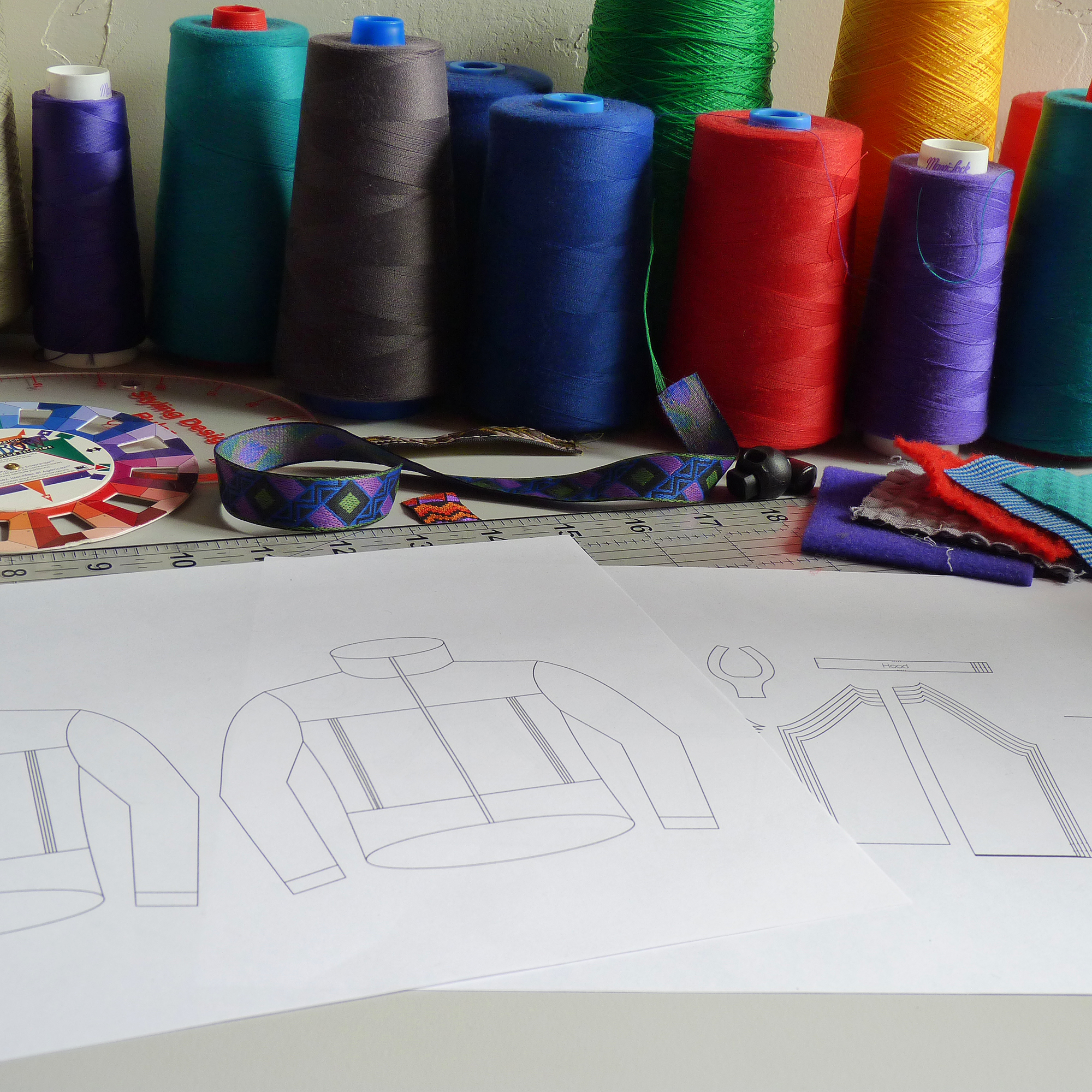 Colorful spools of thread, a fabric swatch, a measuring ruler, a color wheel, and sewing pattern sketches on paper, indicating a sewing or fashion design workspace.