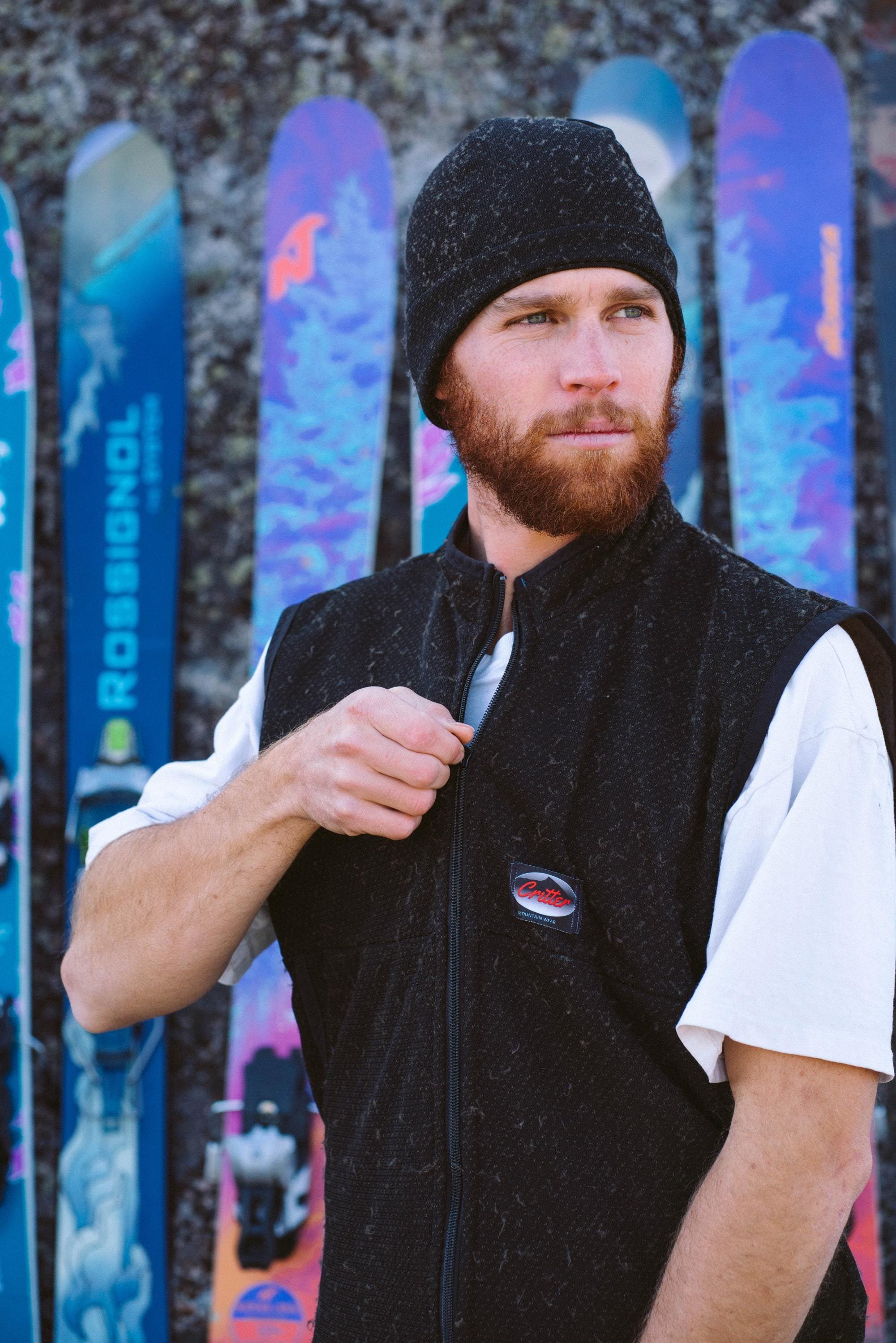 A man is zipping up the front of a Polartec Critter Birthday Suit while standing in front of some skis.