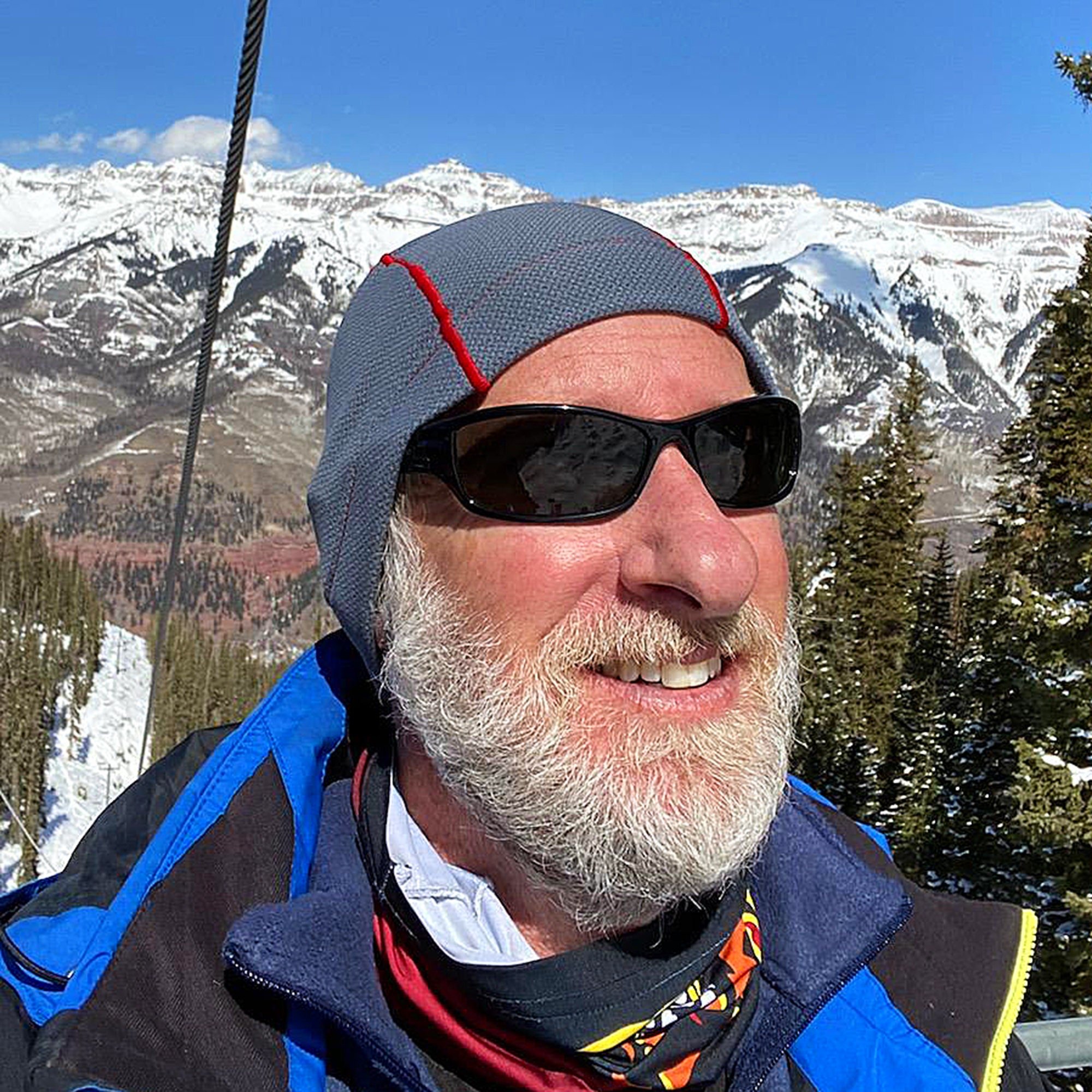A man wearing a grey Polartec hat with a red stripe, black sunglasses, a blue and black jacket, and a black and orange neck gaiter.