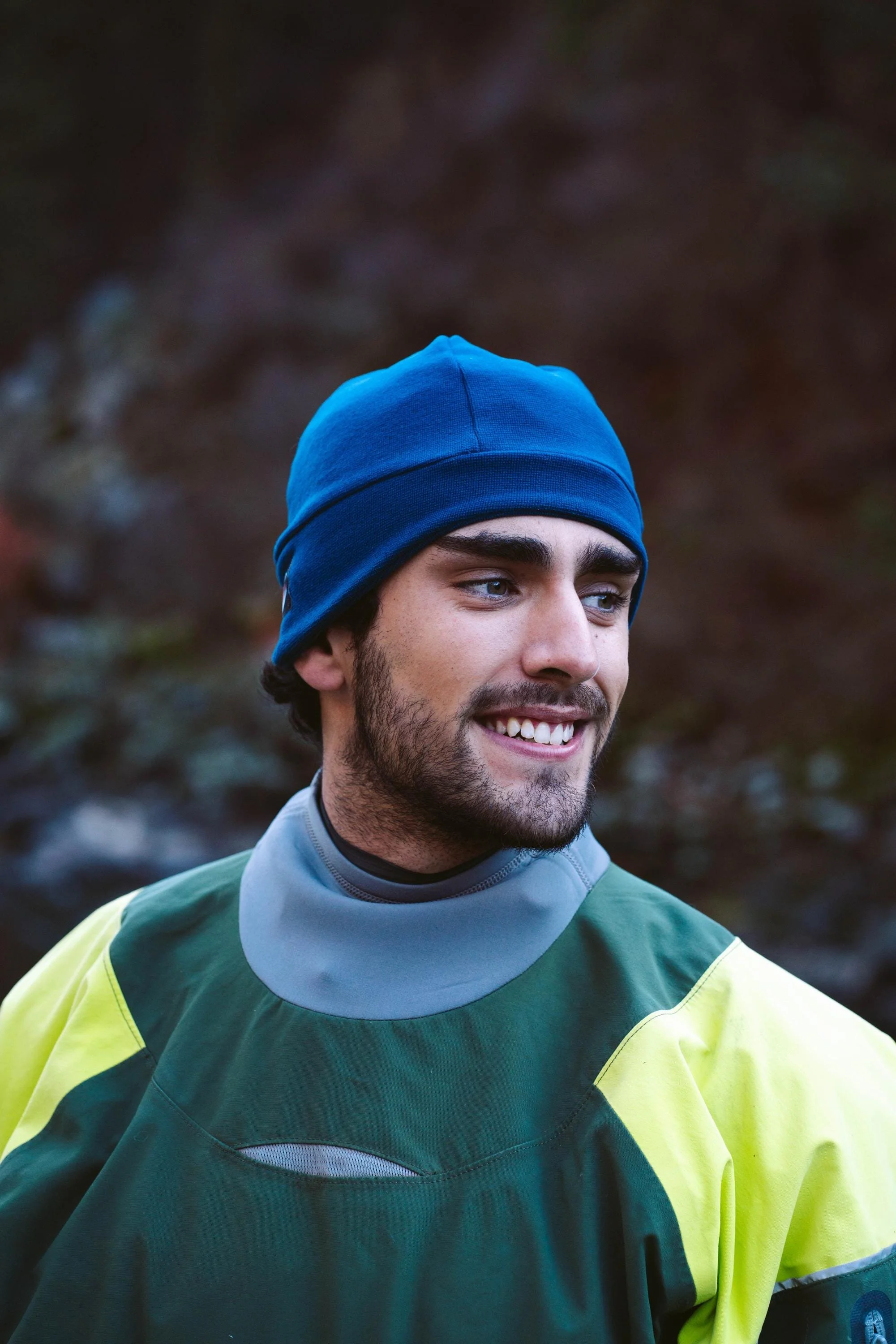 A man smiling wearing a blue fleece hat while standing next to a river.