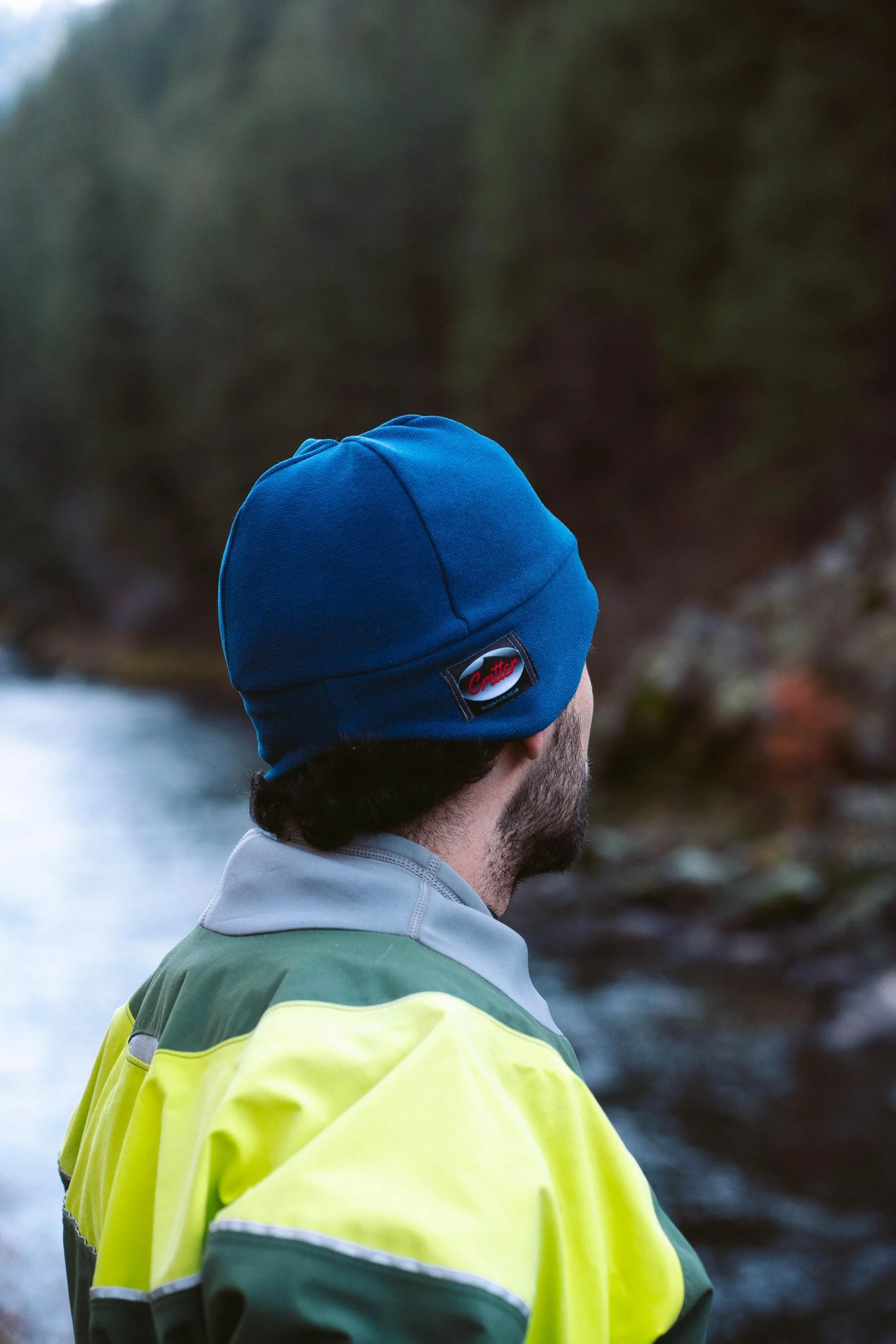 A man wearing a blue beanie and a yellow and green waterproof jacket, standing near a river with a forested background, looking away from the camera.