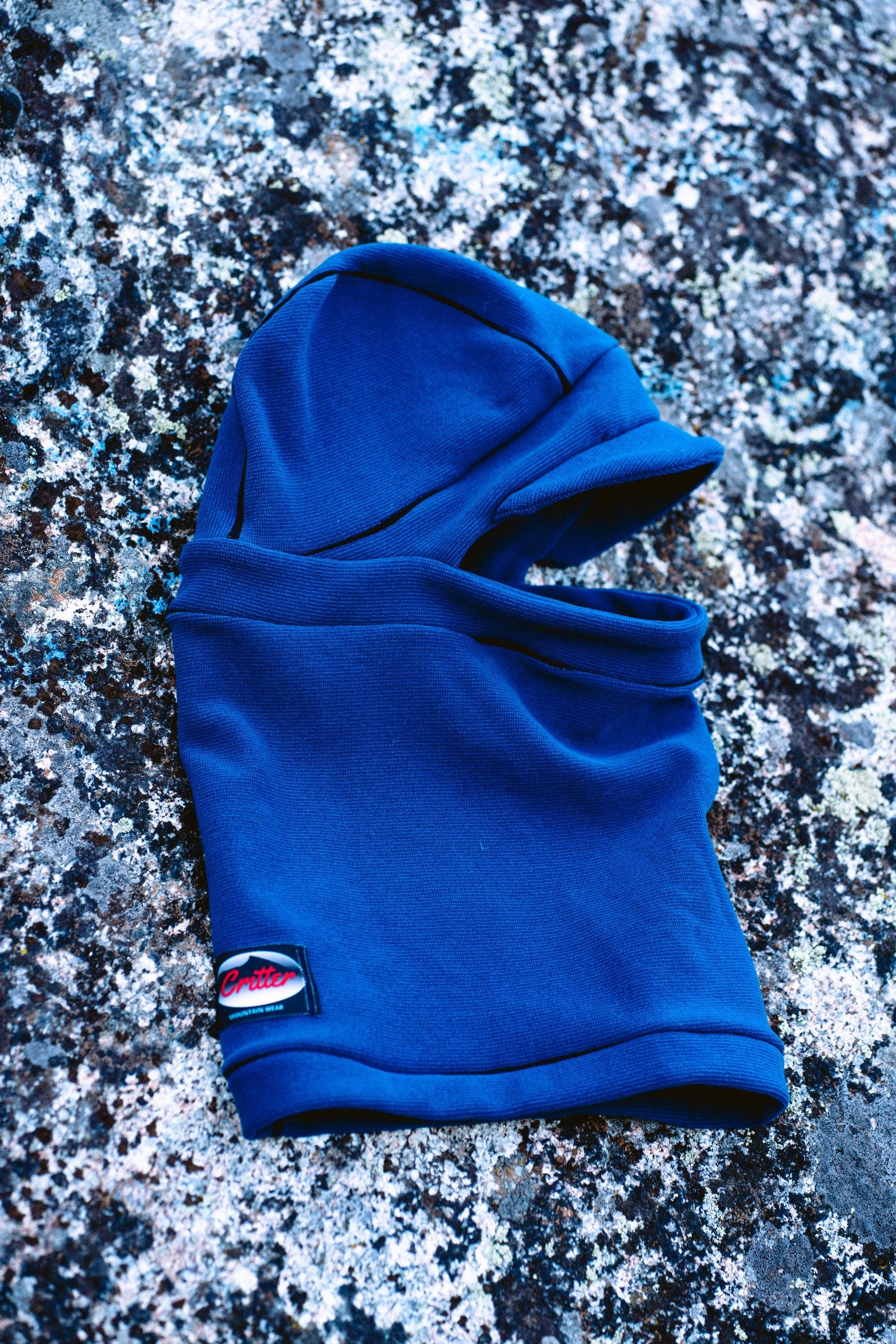 A blue balaclava with blue trim laying on a rock. The balaclava has a fold-down hat with a brim.