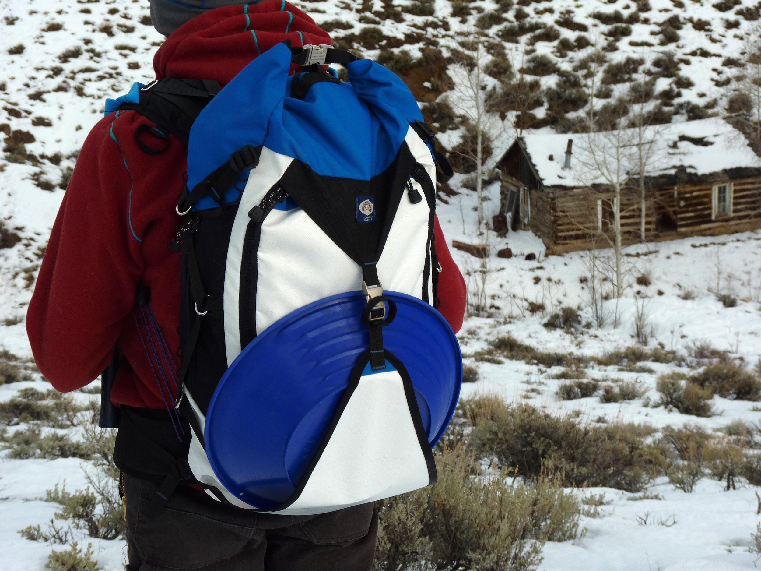 A blue, white, and black backpack with a blue gold pan attached to the front. The backpack has a white strap and a black buckle.