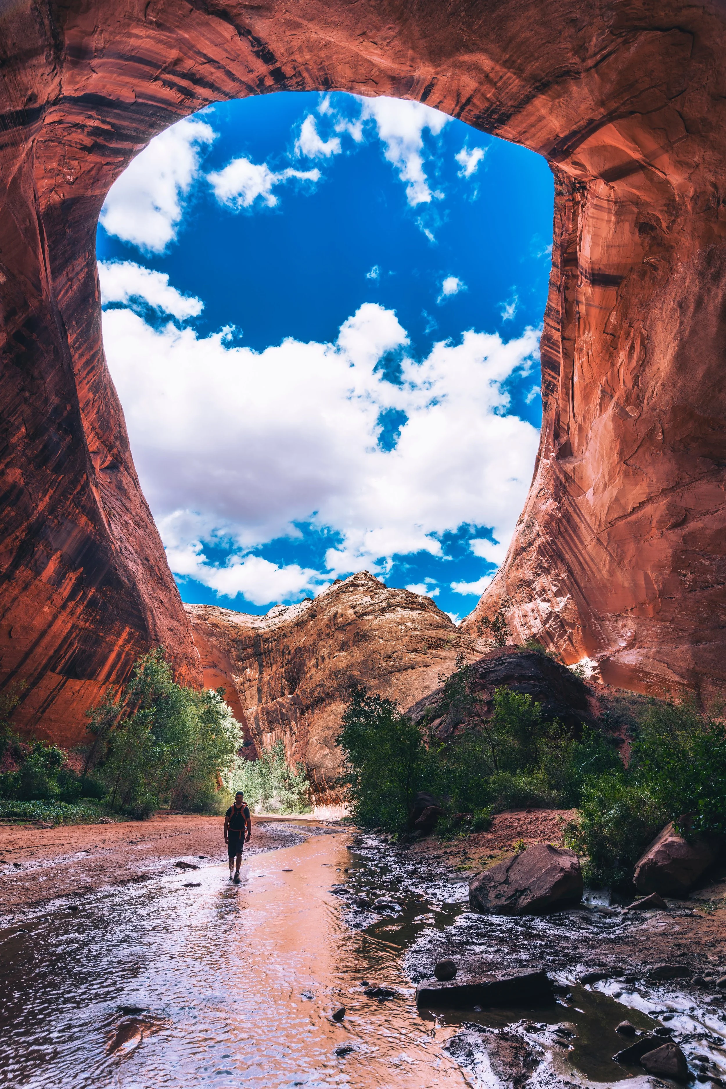 A person hiking in a slot canyon with towering red rock walls and a blue sky with white clouds overhead.