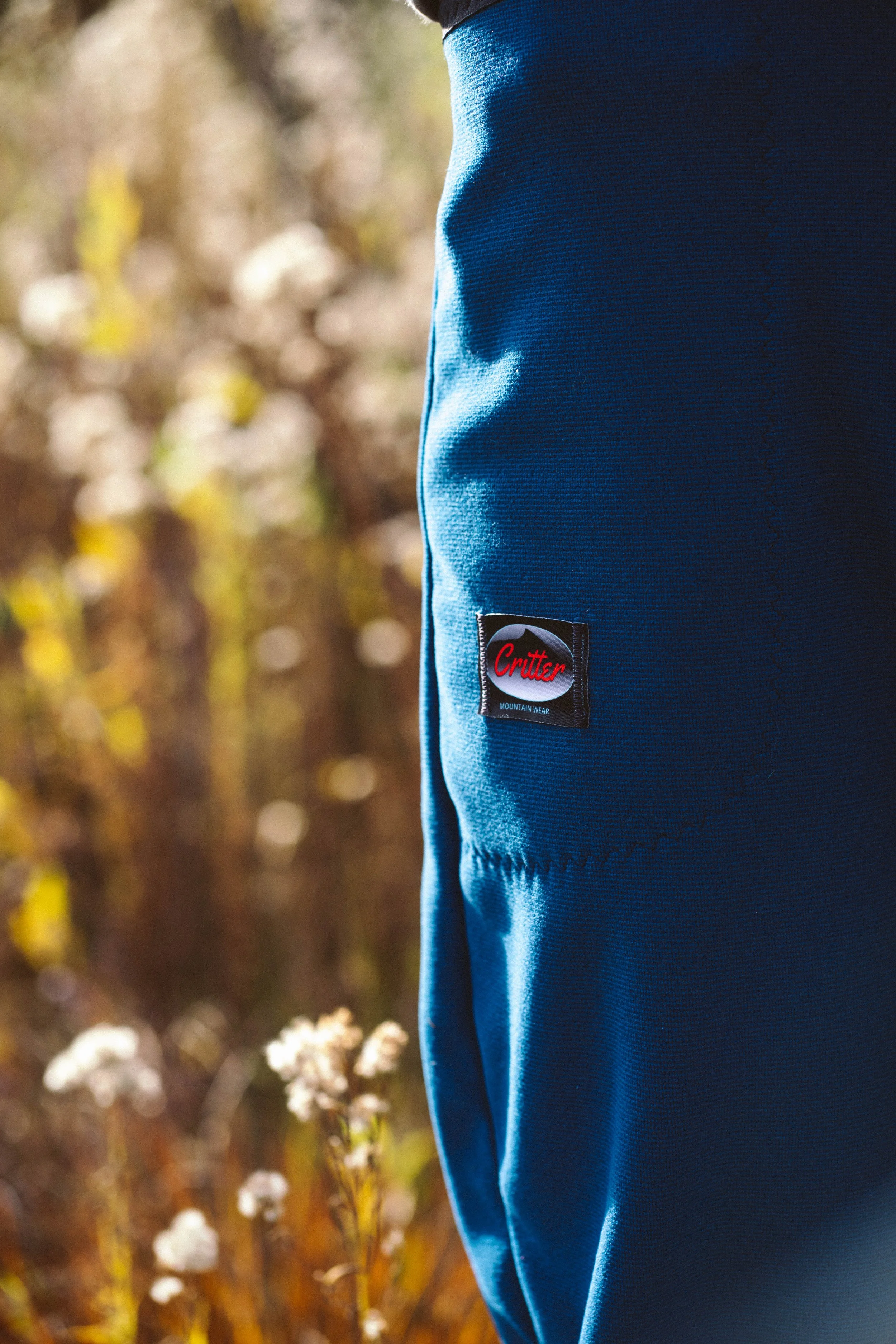 A close-up of a person wearing a blue Critter mountain wear pants outdoors near plants with a blurred background of foliage.