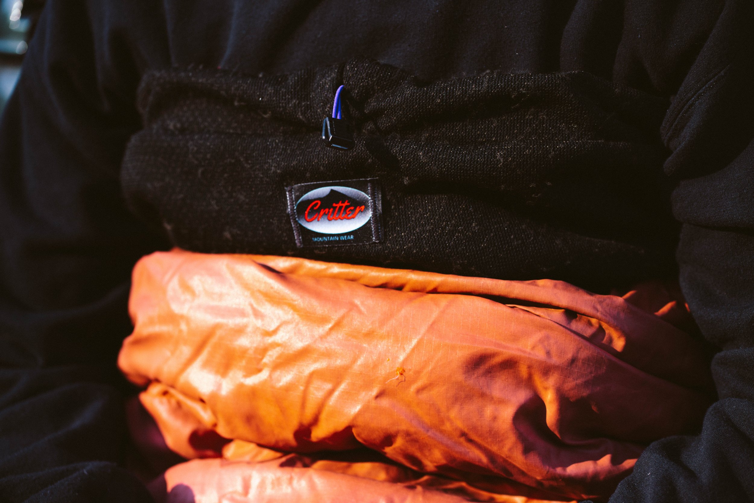 Close-up of a person wearing black outdoor clothing, with an orange sleeping bag or jacket. The clothing has a label that reads 'Critter Mountain Wear.'