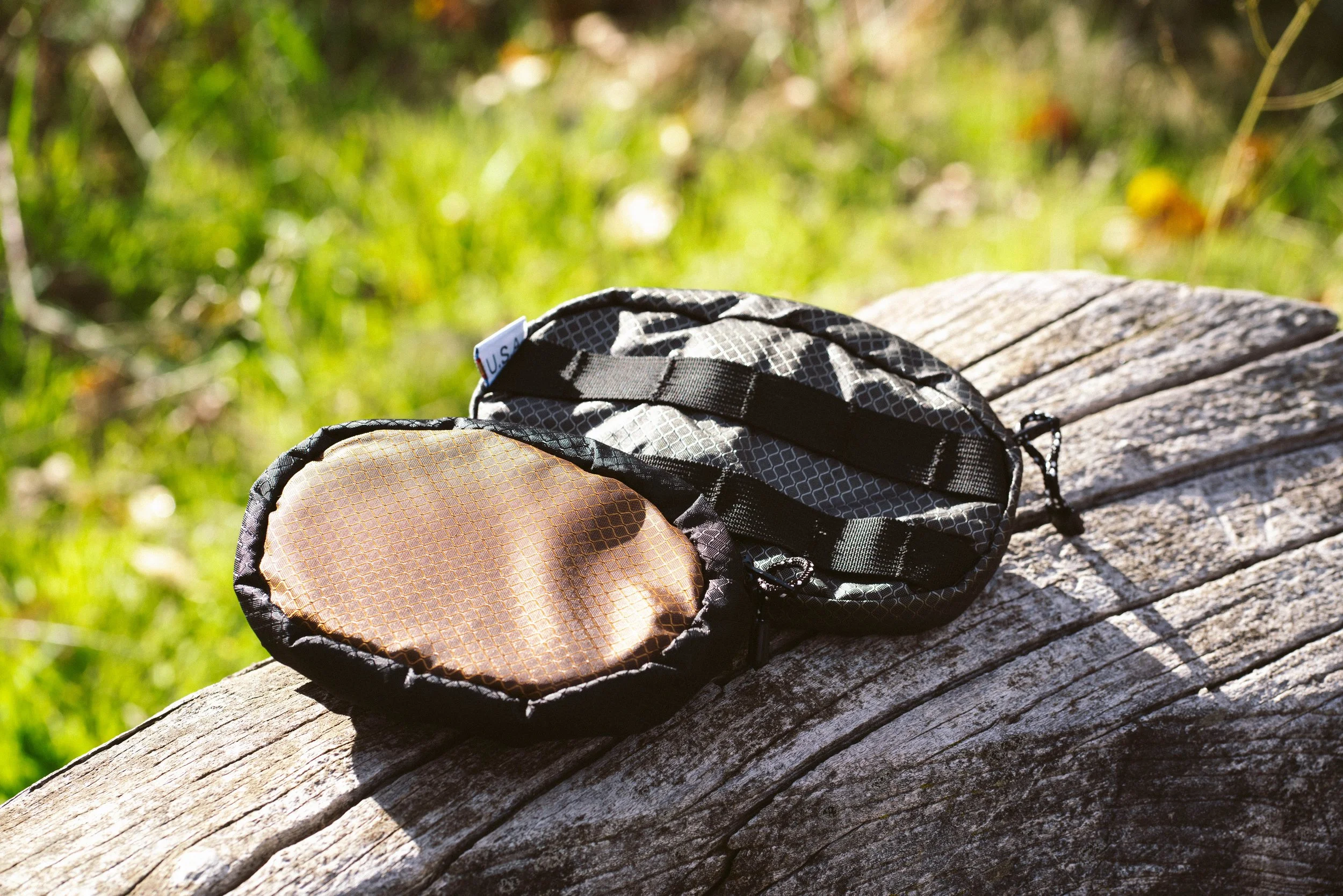 A black and gray patterned camera bag with a brown protective cover on a weathered wooden log outdoors with green grass in the background.