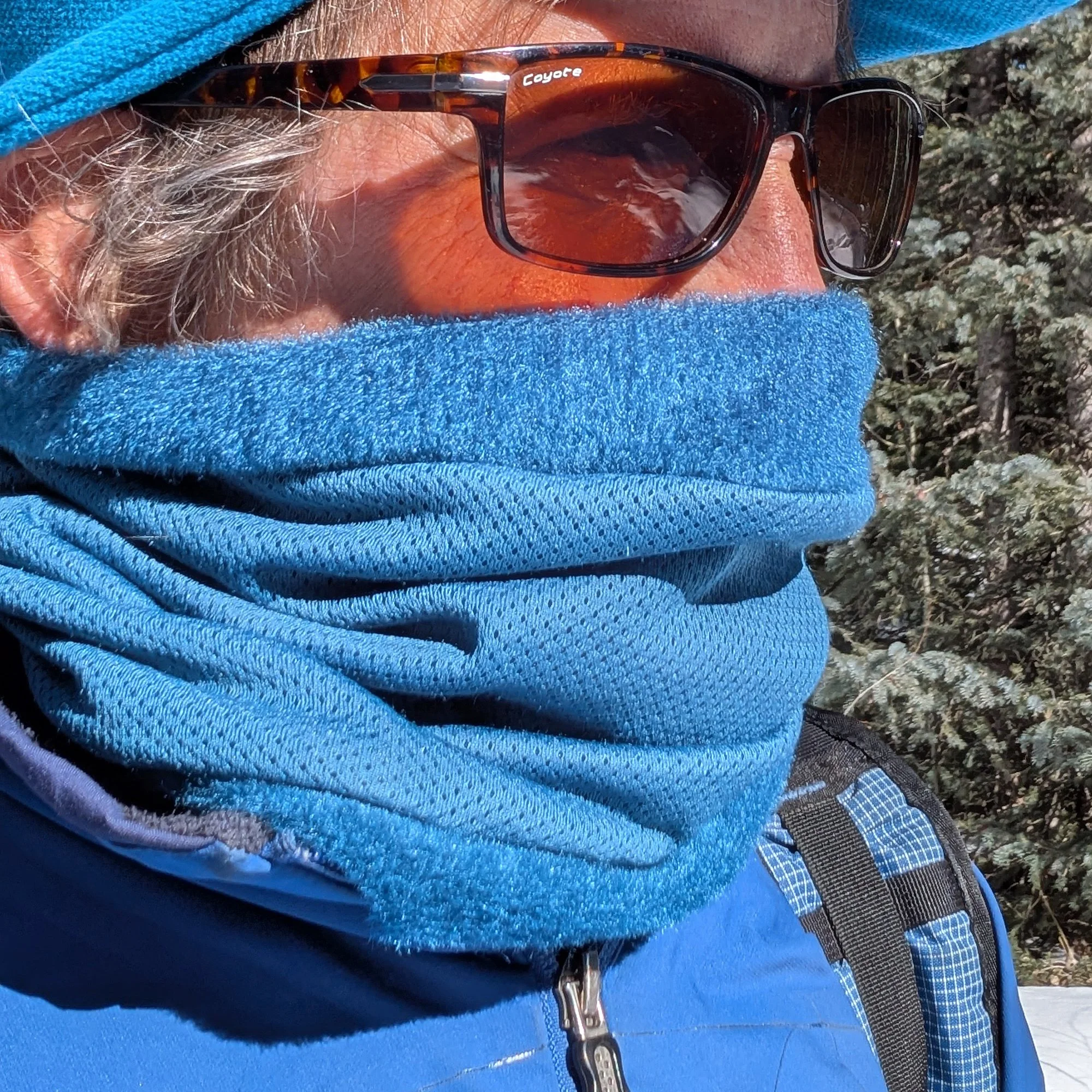 A man wearing a teal green fleece neck gaiter, and sunglasses. The neck gaiter covers the nose and most of the face.