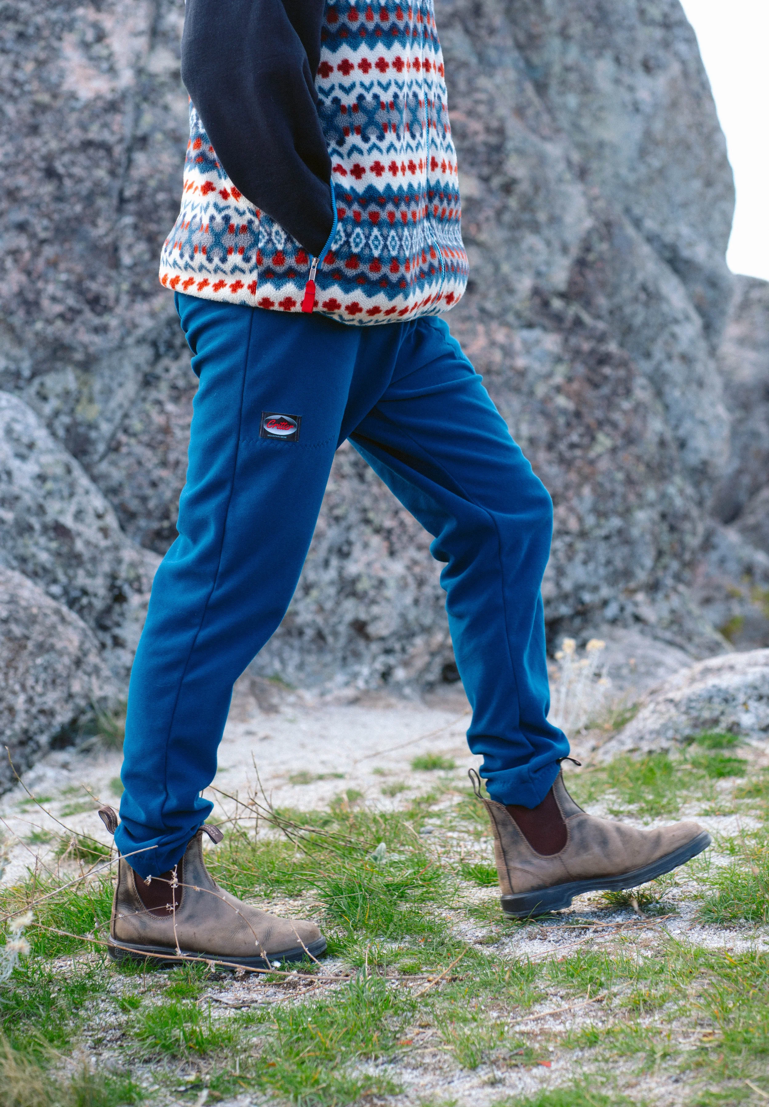 Person standing on rocky terrain wearing beige hiking boots, blue pants, and a patterned sweater with red, white, and blue designs.