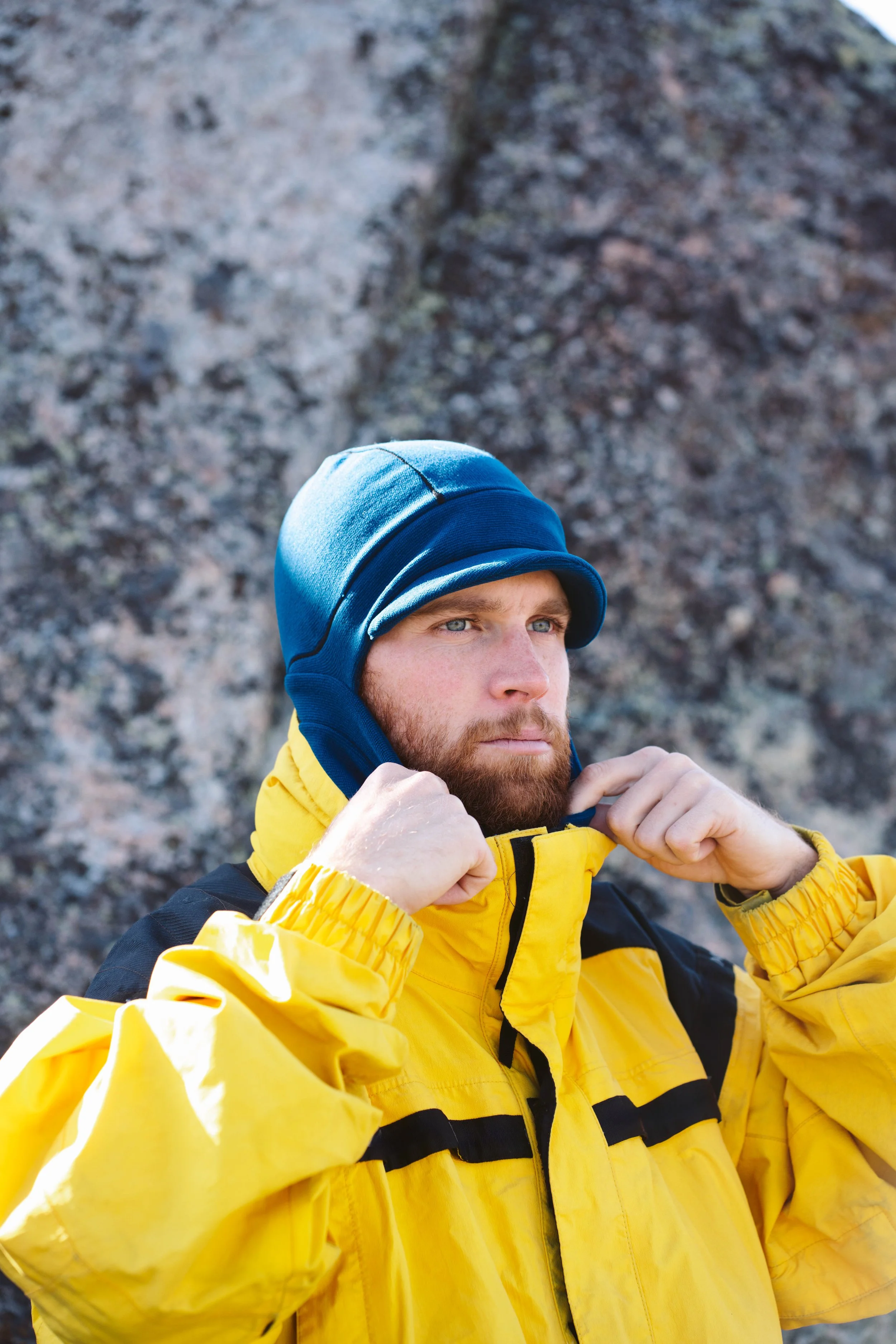 A man wearing blue balaclava with blue trim. The balaclava has a fold-down hat with a brim. The man is pulling up the collar of the balaclava.