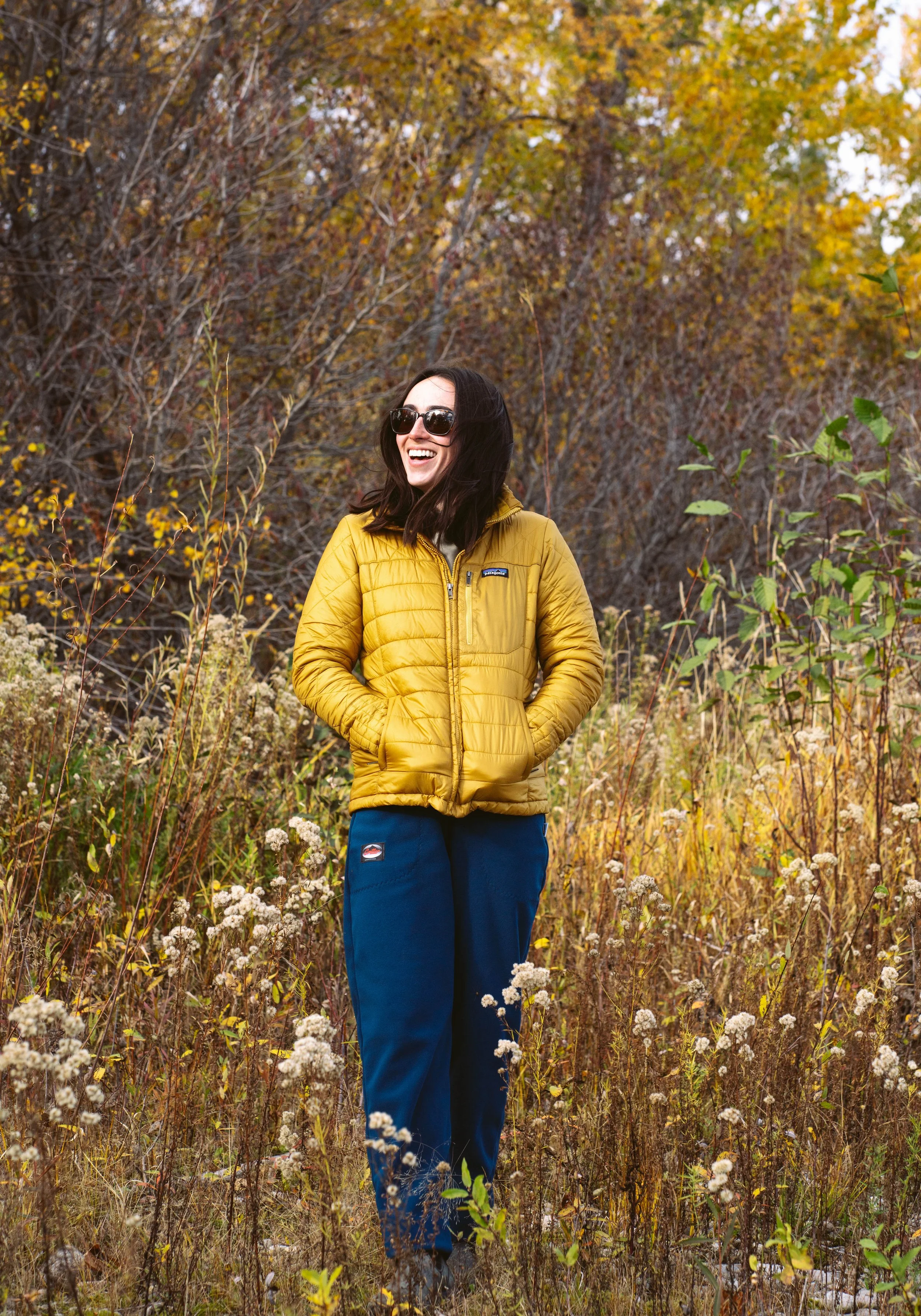 A woman wearing sunglasses, a yellow puffy jacket, and blue pants stands in an autumnal field with trees and shrubs in the background, smiling and looking to the side.