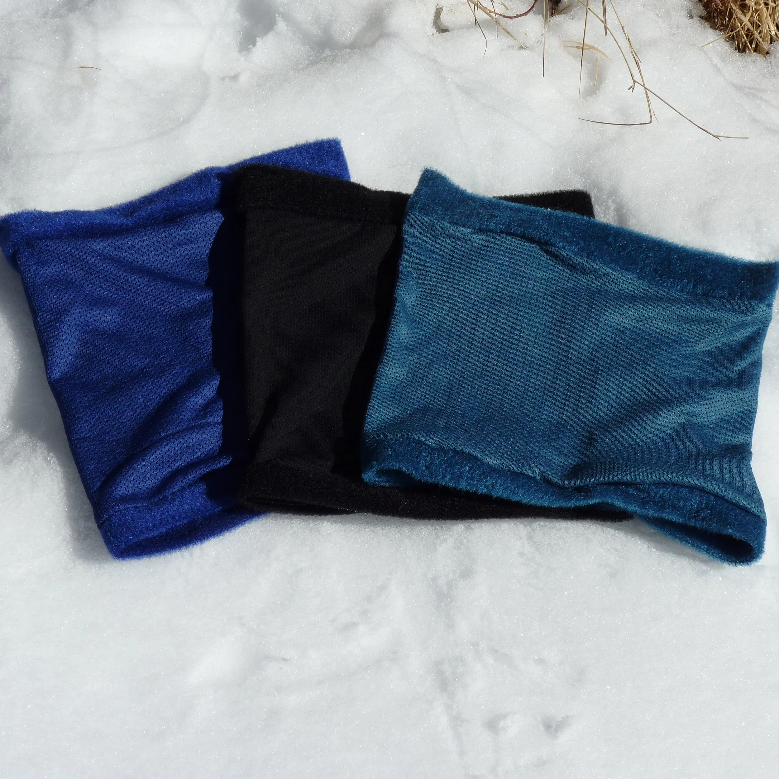 Three fleece neck gaiters laying on snow. One blue neck gaiter. One black neck gaiter. One teal neck gaiter.