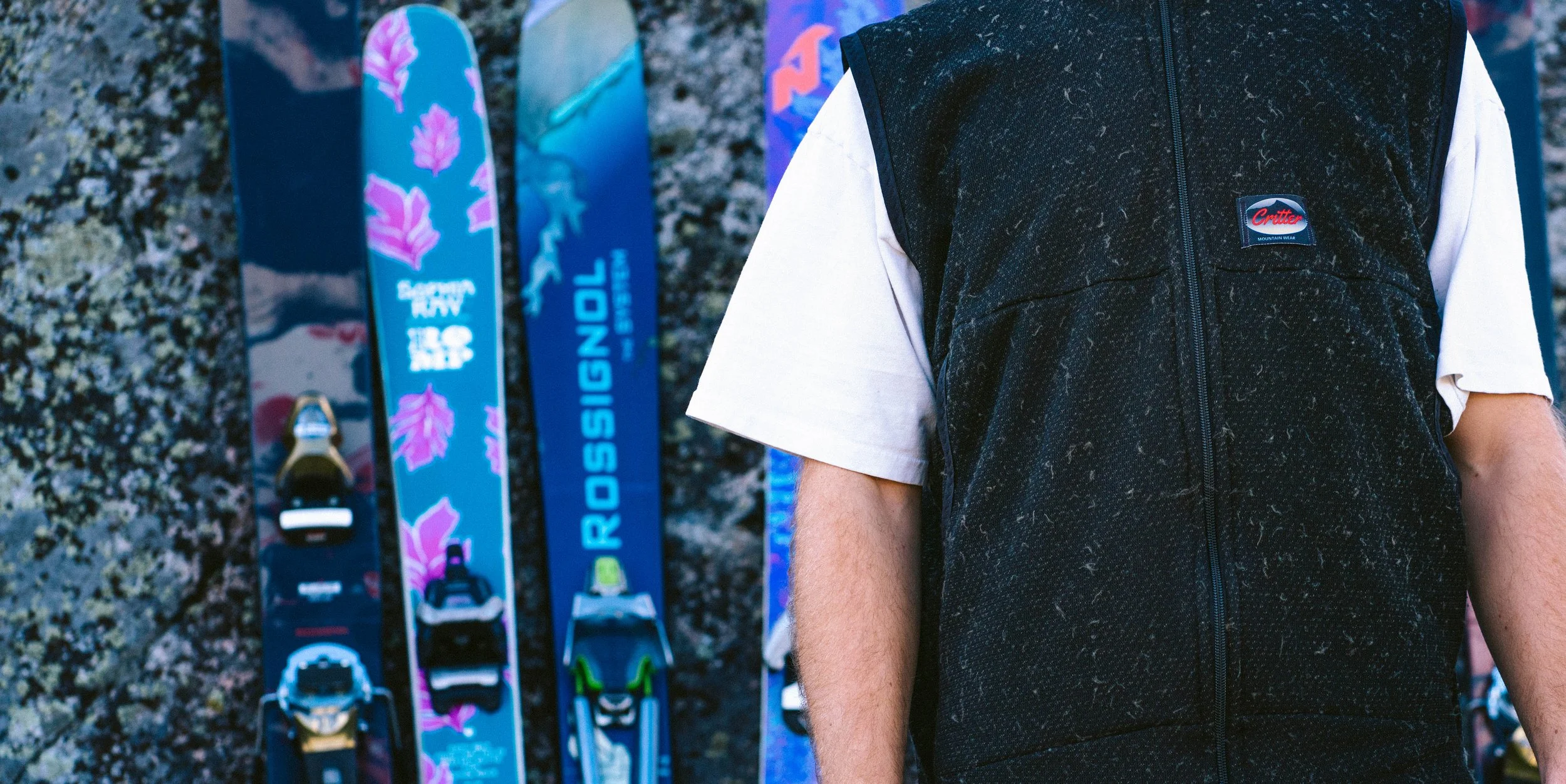 Person wearing a black vest standing outdoors next to a wall of colorful skis with brands like Rossignol and look bindings.