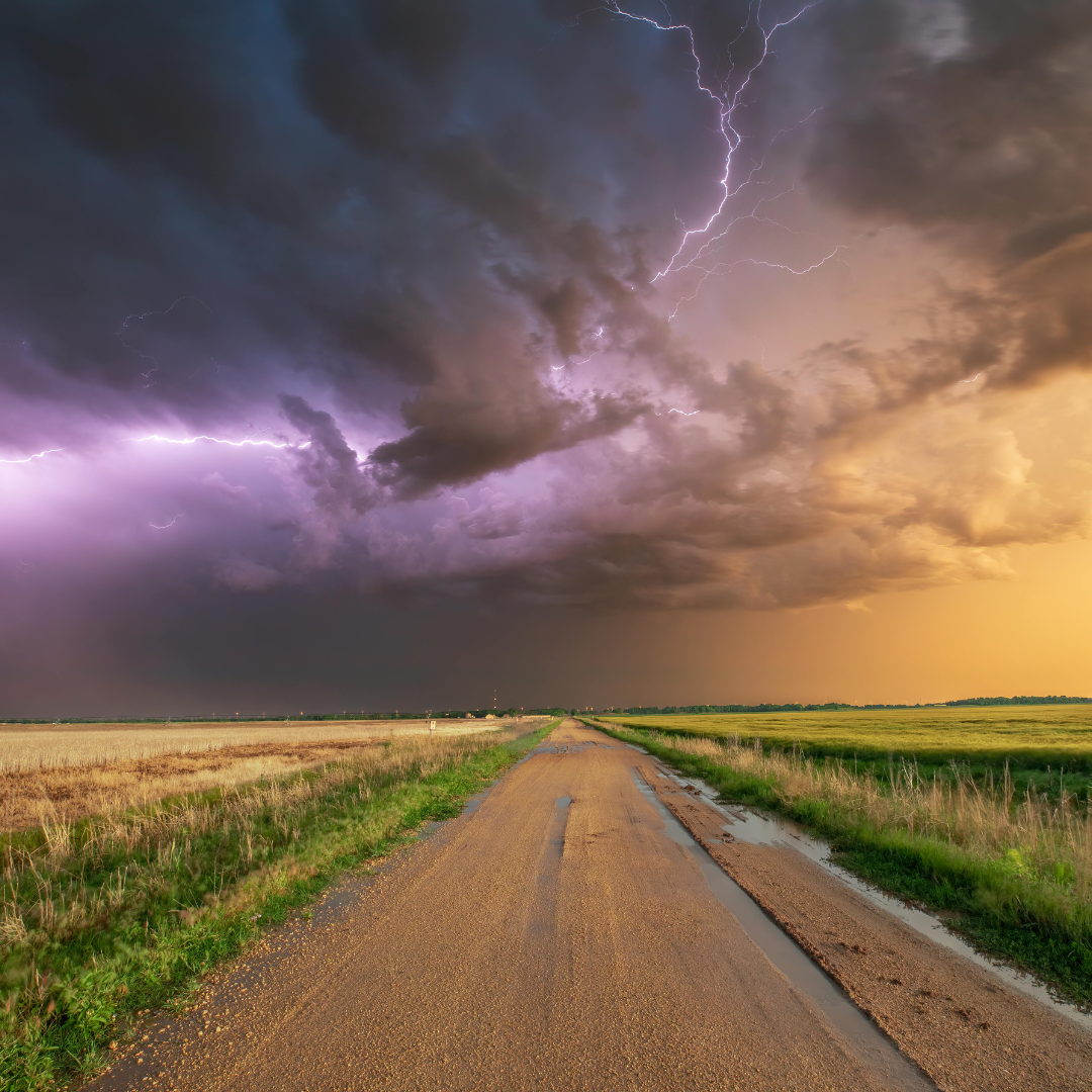 Stormy sky with lightning over a rural dirt road flanked by fields at sunset or sunrise.