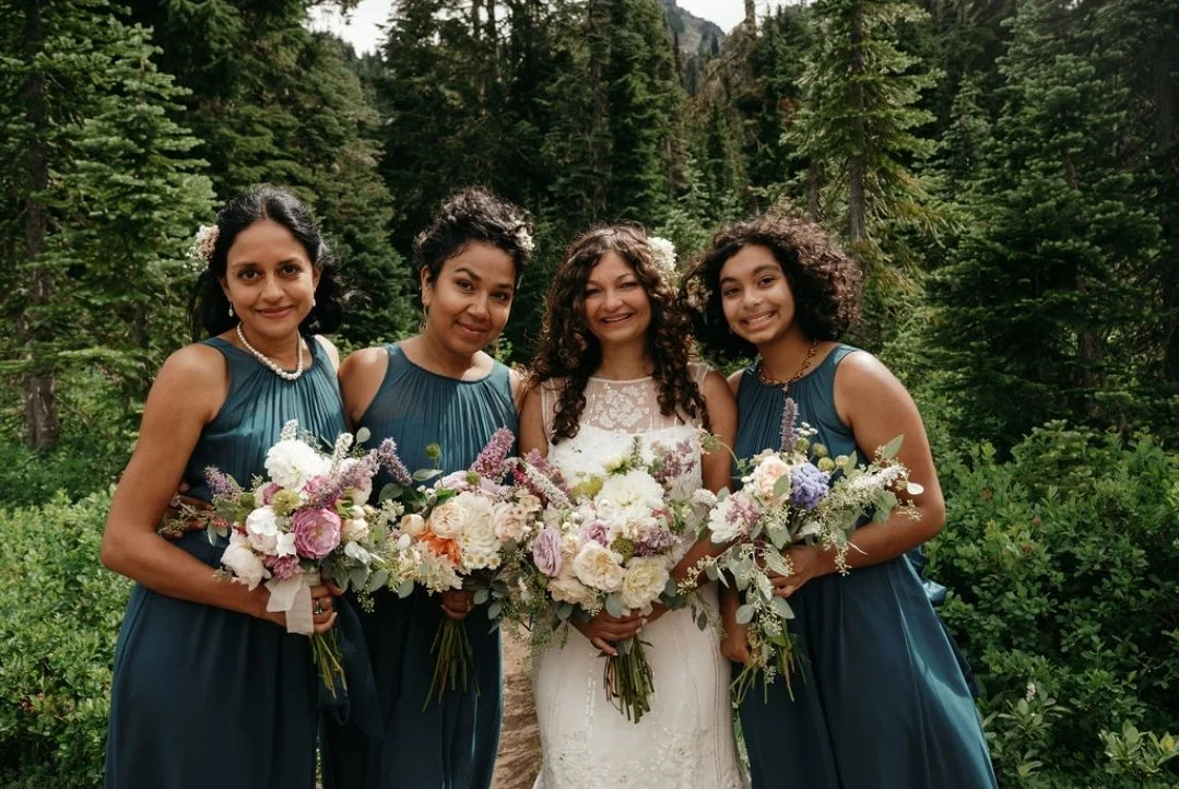Four women in formal dresses holding bouquets of flowers outdoors among trees, at a wedding.