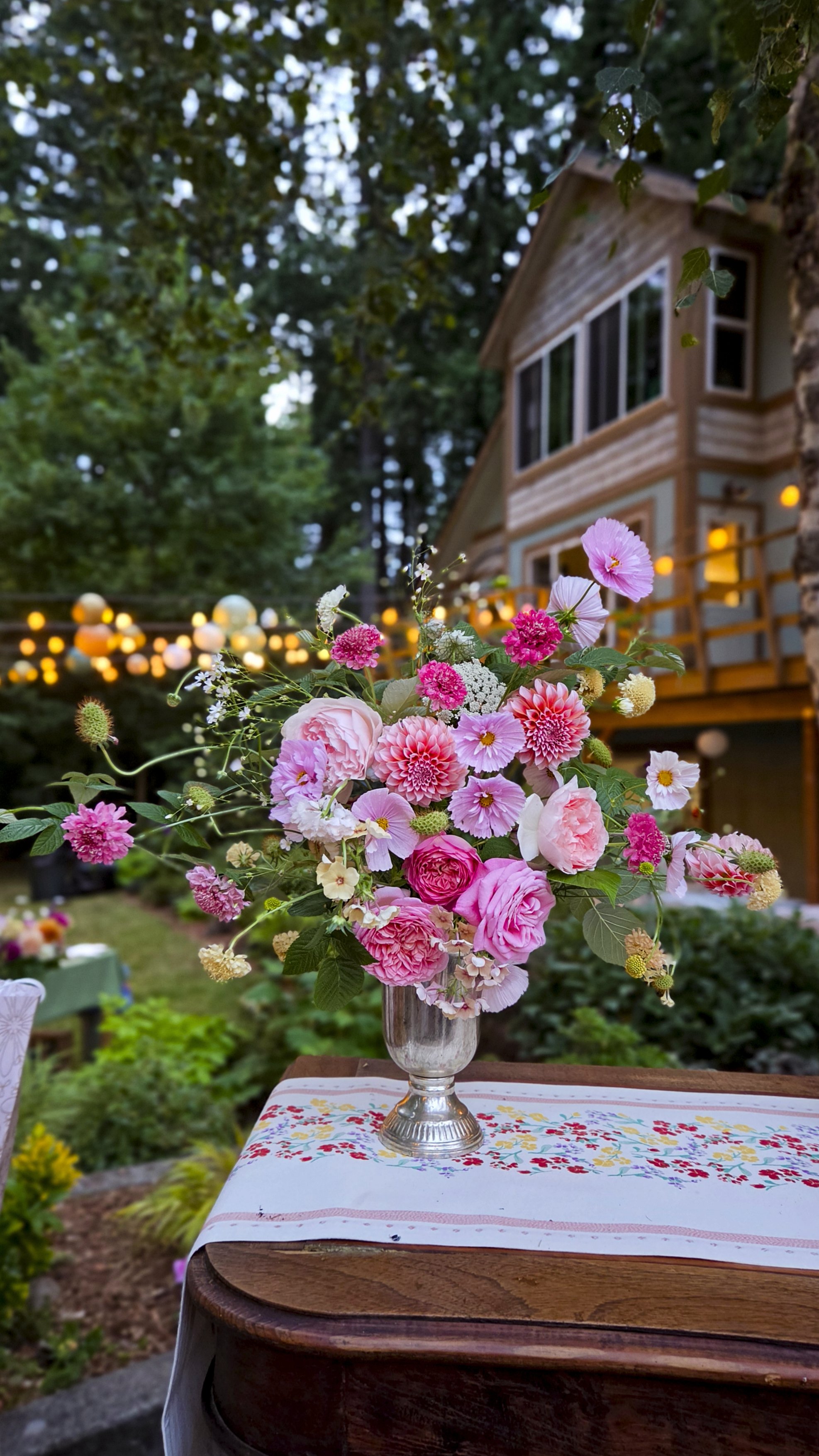 A silver vase filled with pink, white, and cream flowers, placed on a table with a colorful embroidered tablecloth, outdoors in a garden with blurred background, house, trees, and string lights.