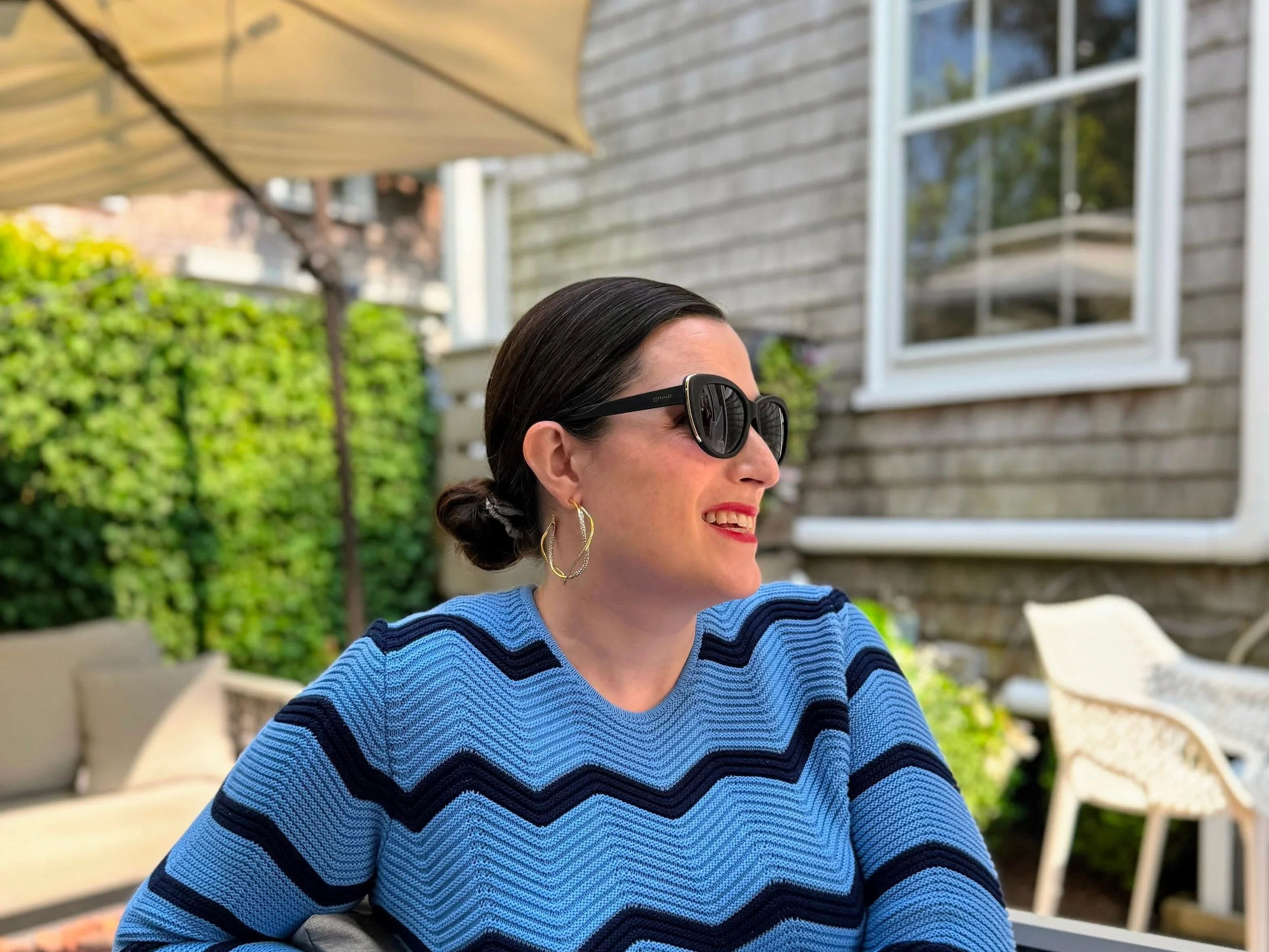 A woman with dark hair pulled back, wearing large black sunglasses, gold earrings, and a blue striped sweater, sits outside in a garden patio area smiling. There is a beige umbrella, green bushes, a house with gray wooden siding, and outdoor furniture visible in the background.