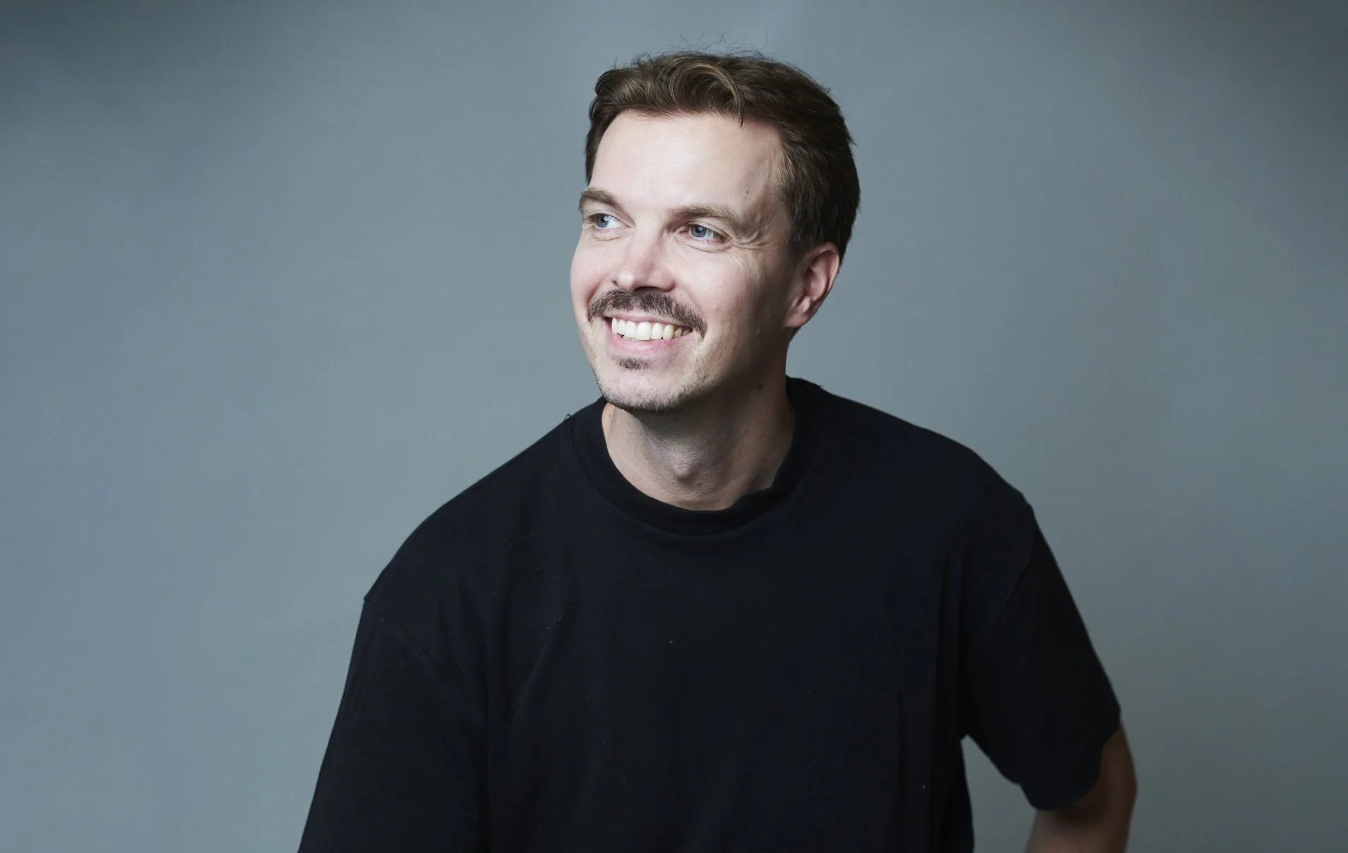A smiling young man with brown hair and a mustache, wearing a black t-shirt, looking to the side against a plain grey background.