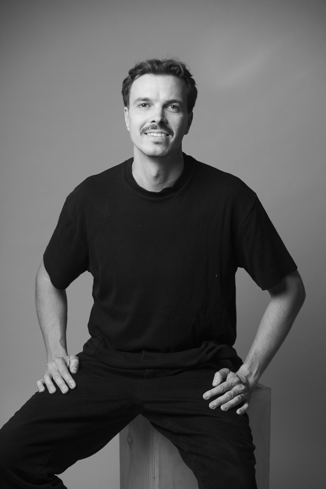 Black and white portrait of a man sitting on a wooden stool, smiling, wearing a black T-shirt and pants, with a neutral background.